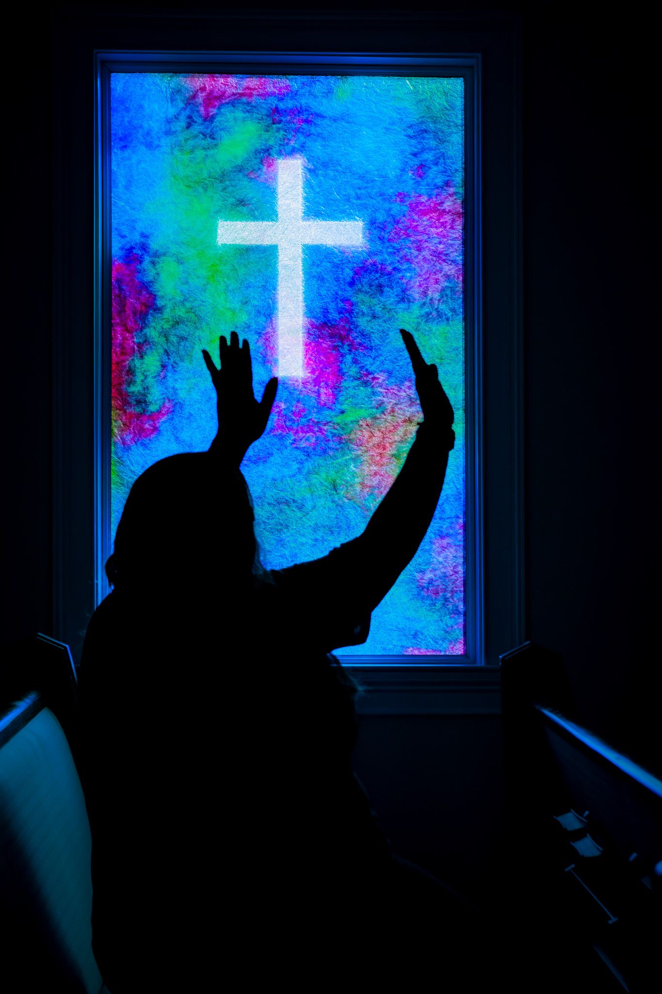 Silhouette of a person with raised hands in worship before a colorful stained glass window with a glowing white cross.