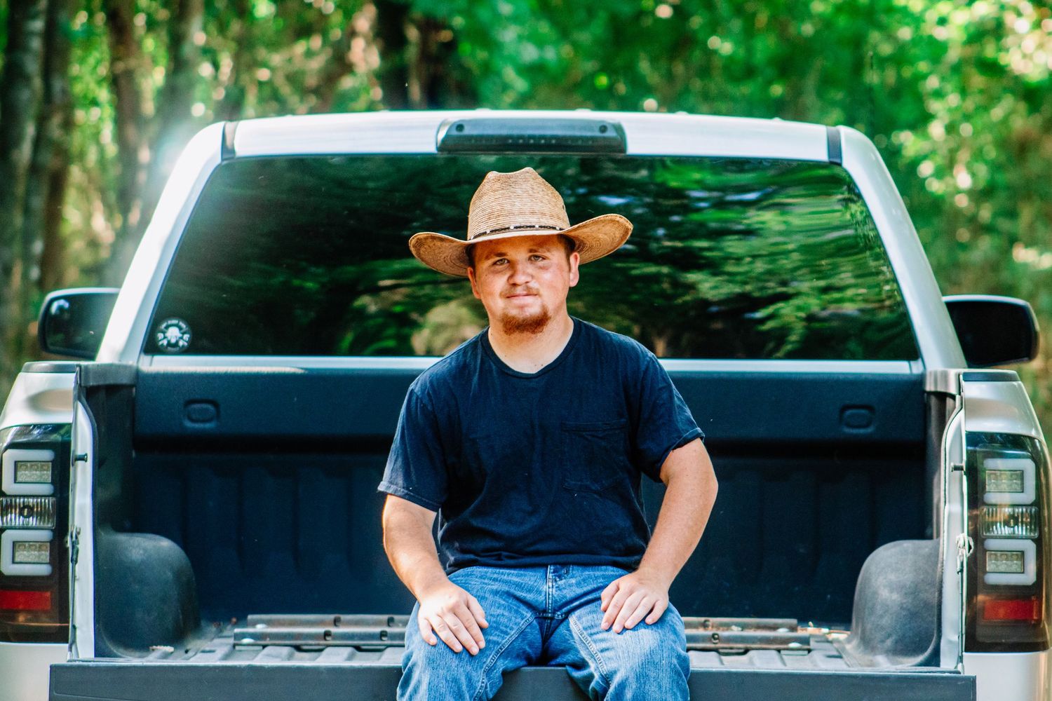 Young man in black t-shirt and cowboy hat sitting in the bed of a pickup truck in a wooded area.