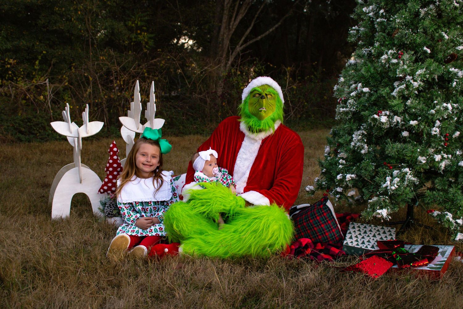Girl and baby with Grinch character sitting by Christmas tree with white reindeer decorations.