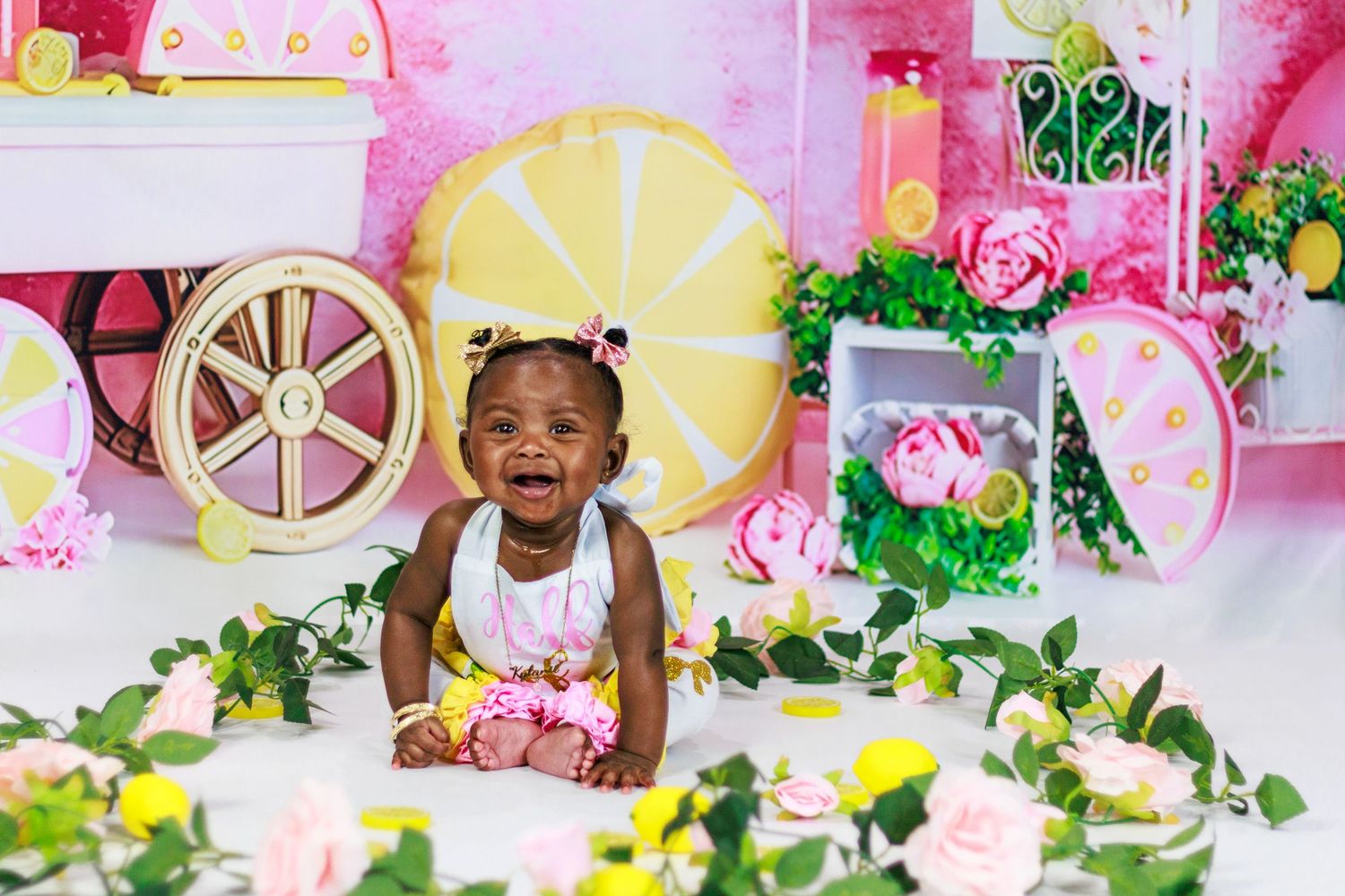 Laughing baby girl lying on her tummy surrounded by flowers and greenery in a lemon-themed photo shoot setting.