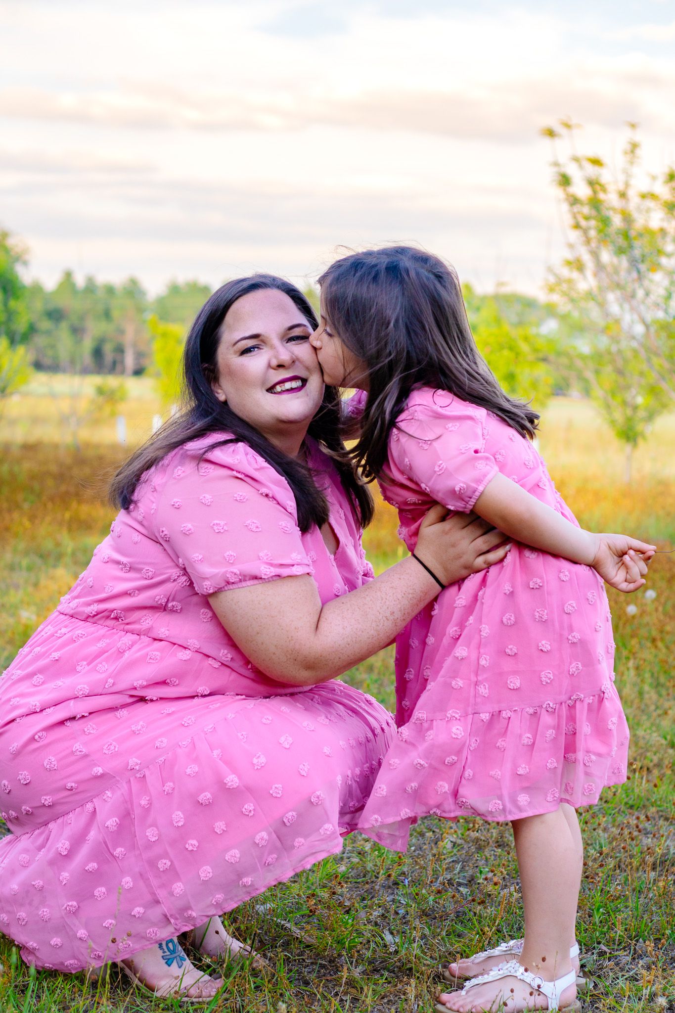 A young girl in a pink dress kisses her smiling mother, both wearing matching pink outfits in a sunny outdoor field.