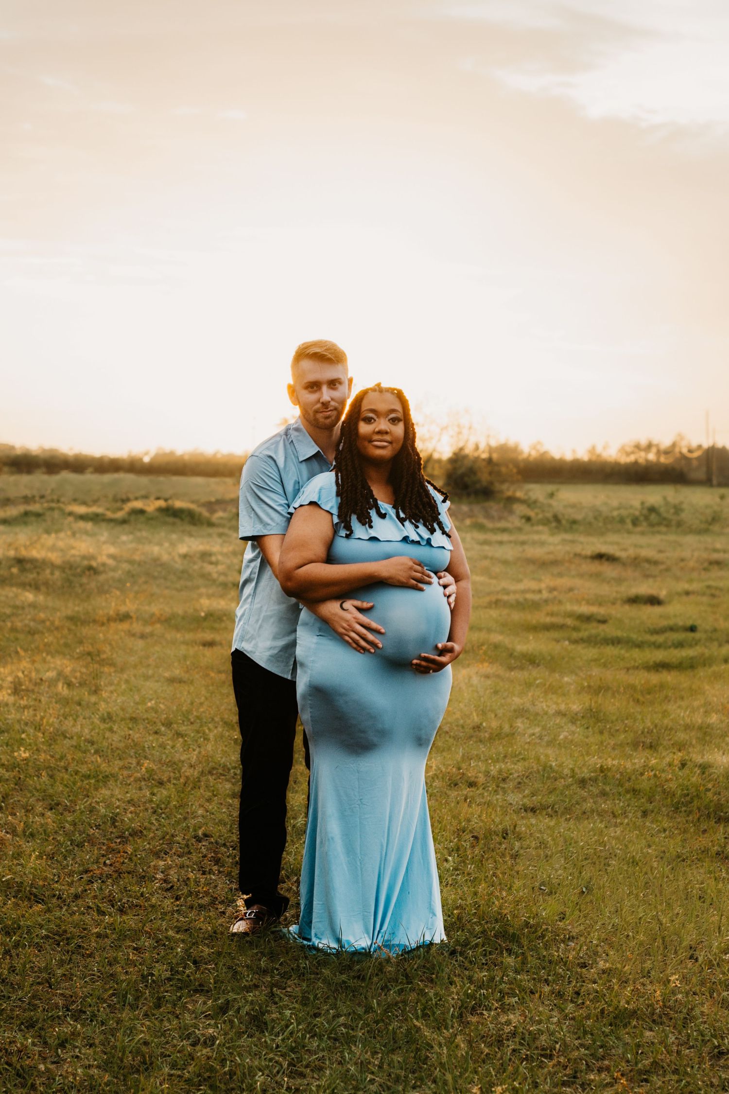 Man tenderly embraces pregnant partner in blue maternity gown during golden hour outdoor photo session.