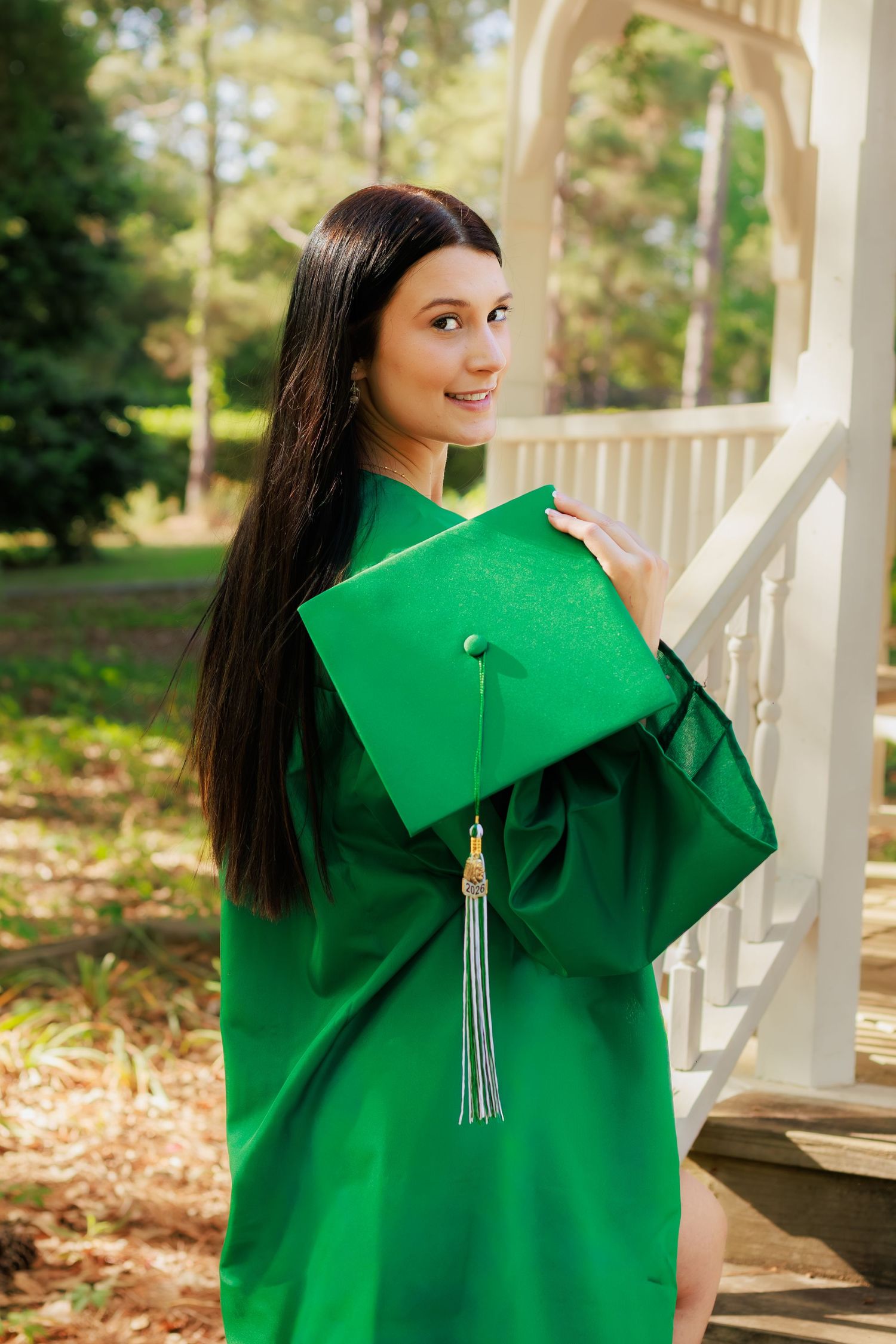 Young woman in green graduation cap and gown smiling outdoors near white porch railing surrounded by trees.