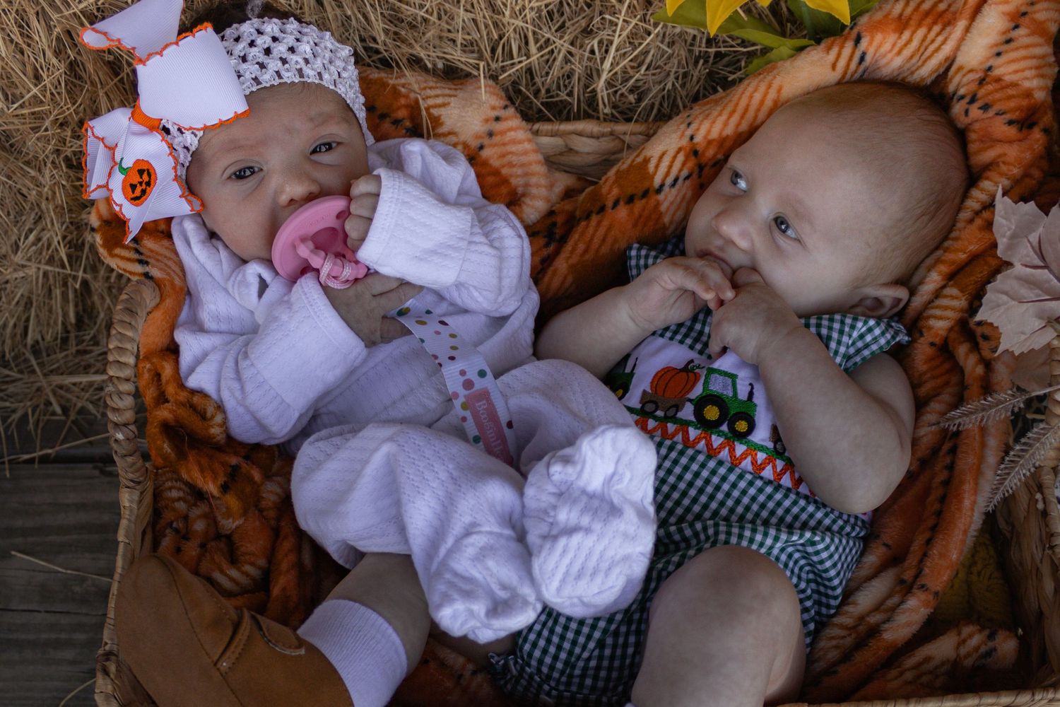 Two newborn babies in fall outfits rest together in a wicker basket with orange blanket.