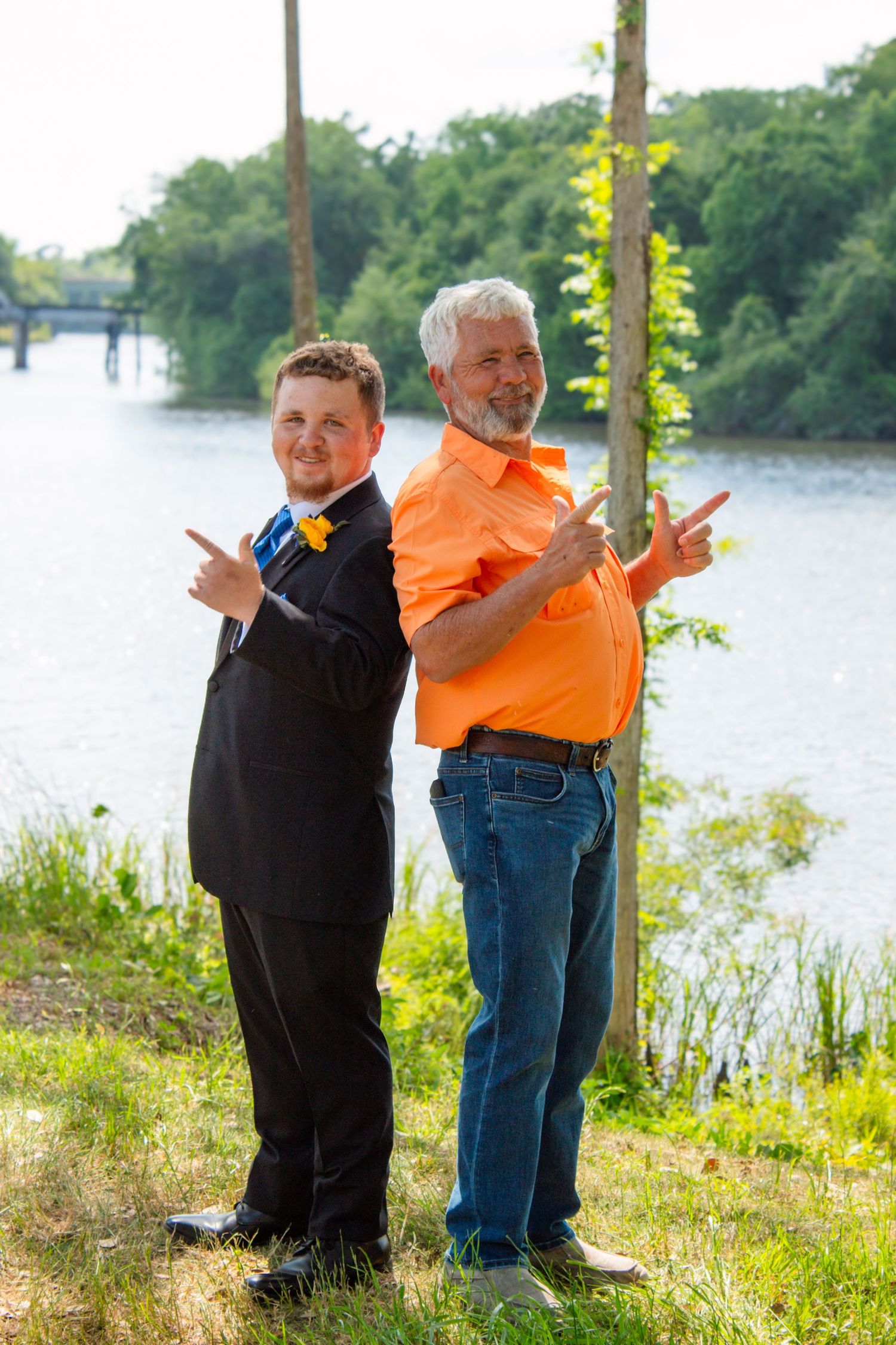 Young man in black prom tuxedo and older man in orange shirt pose back-to-back near riverside.