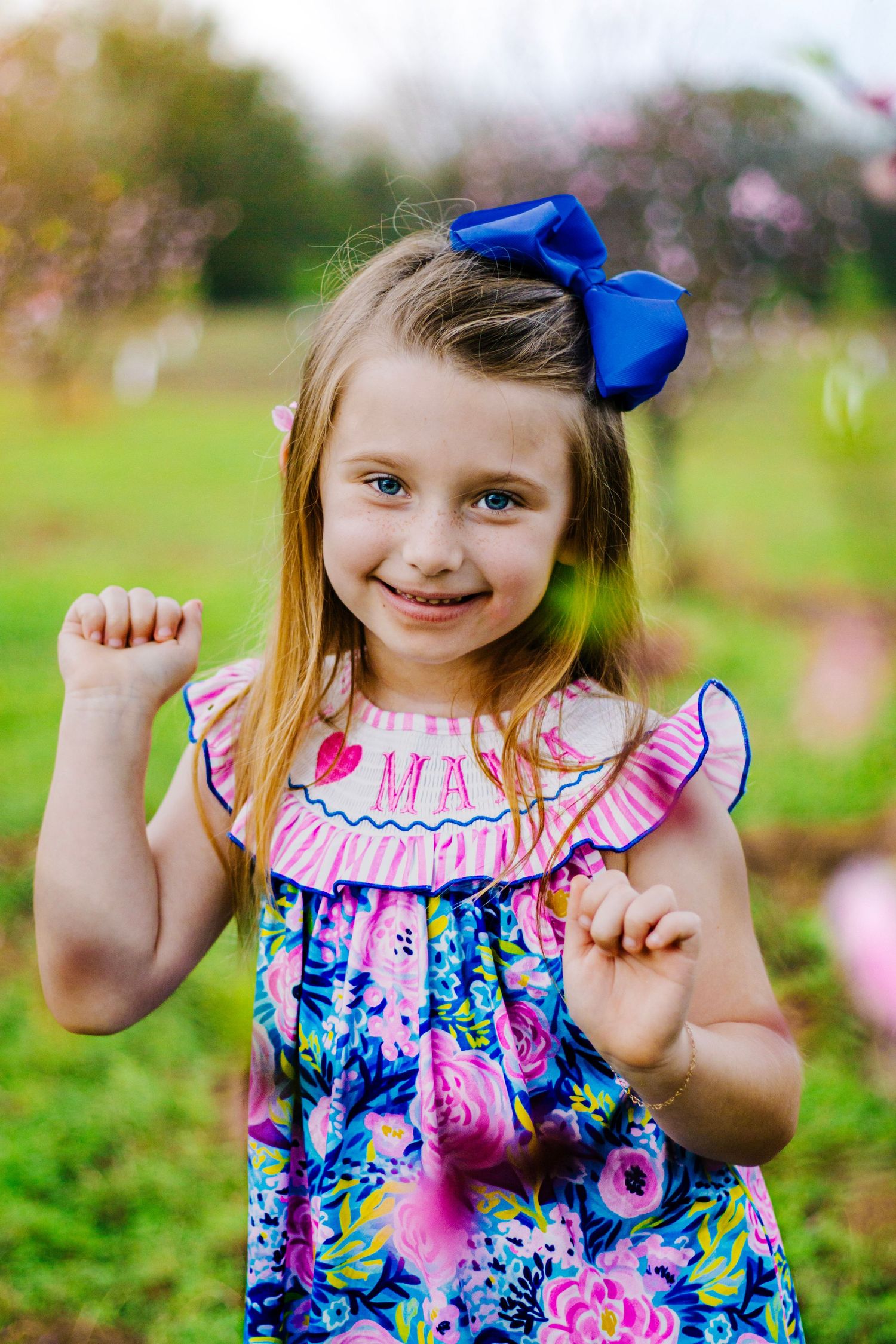Happy young girl in colorful floral dress with blue bow flexing arms among spring blooming trees.