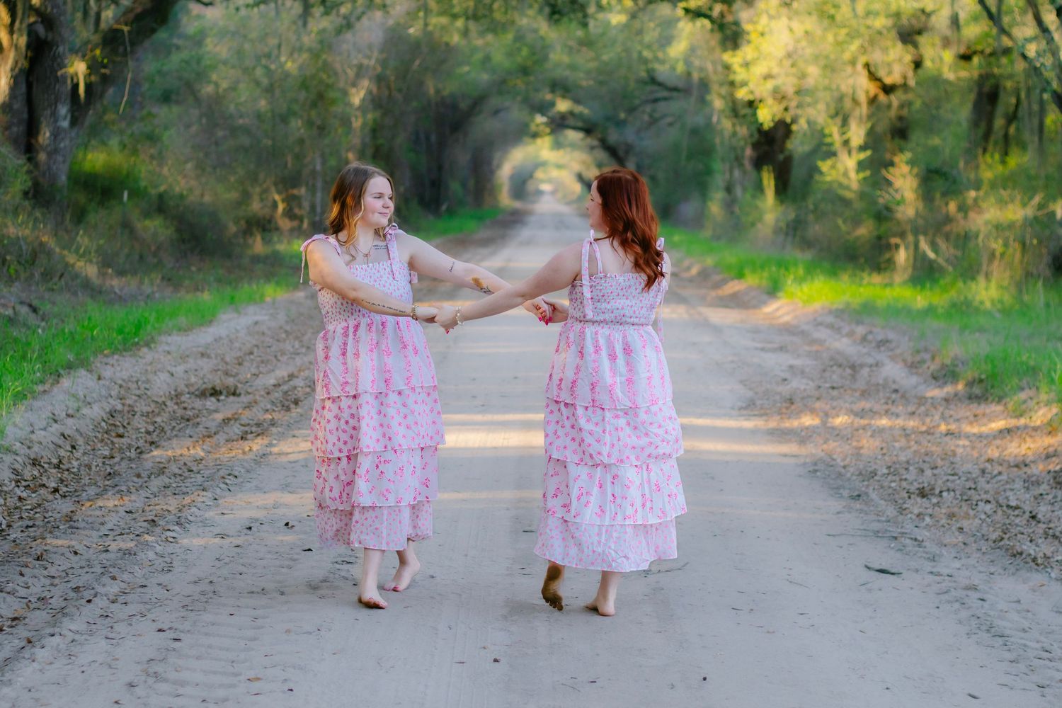 Two women in matching pink floral dresses hold hands and twirl on a scenic tree-lined dirt road.
