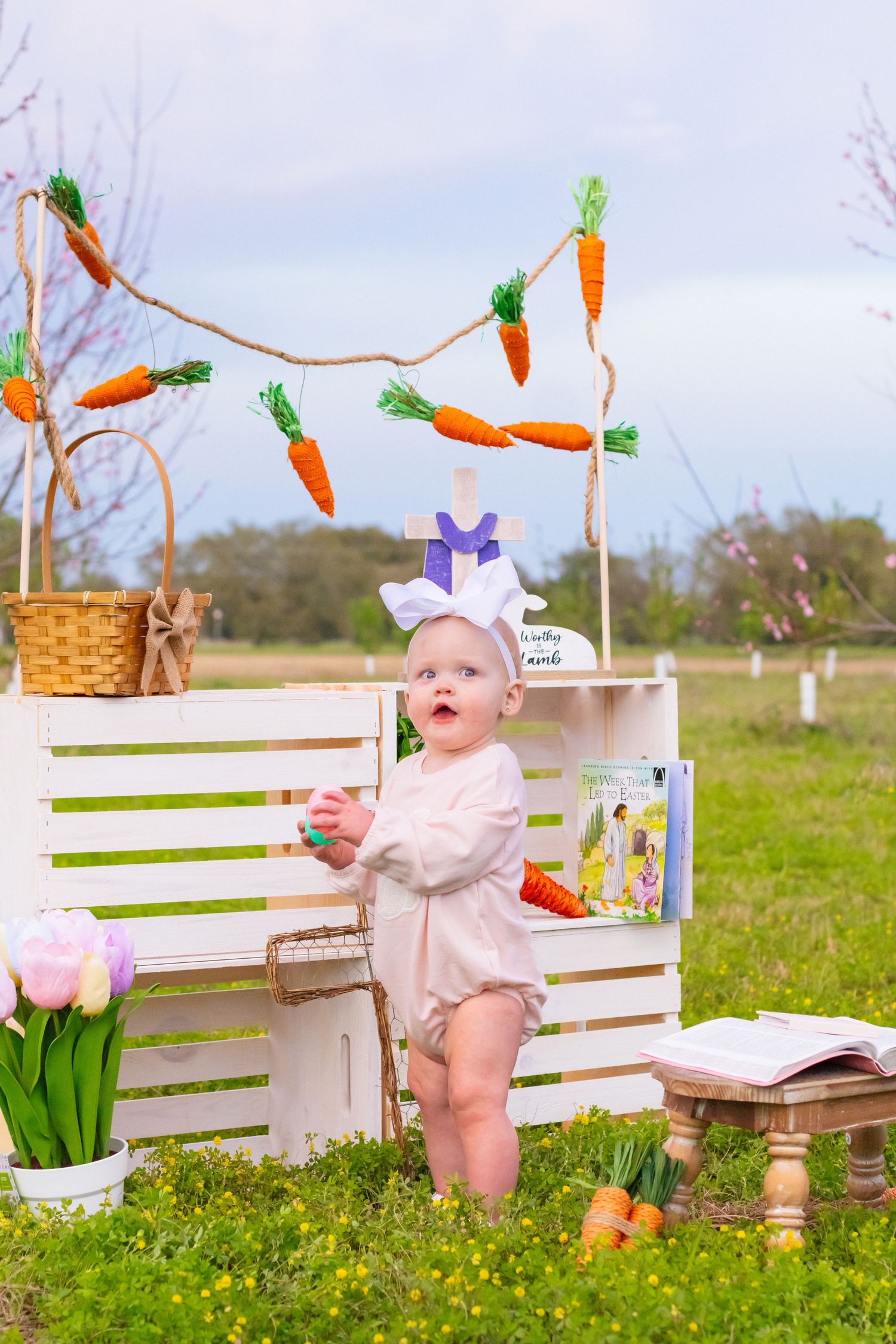 A baby in bunny ears stands by white crates decorated with carrot garland in an Easter outdoor setting.