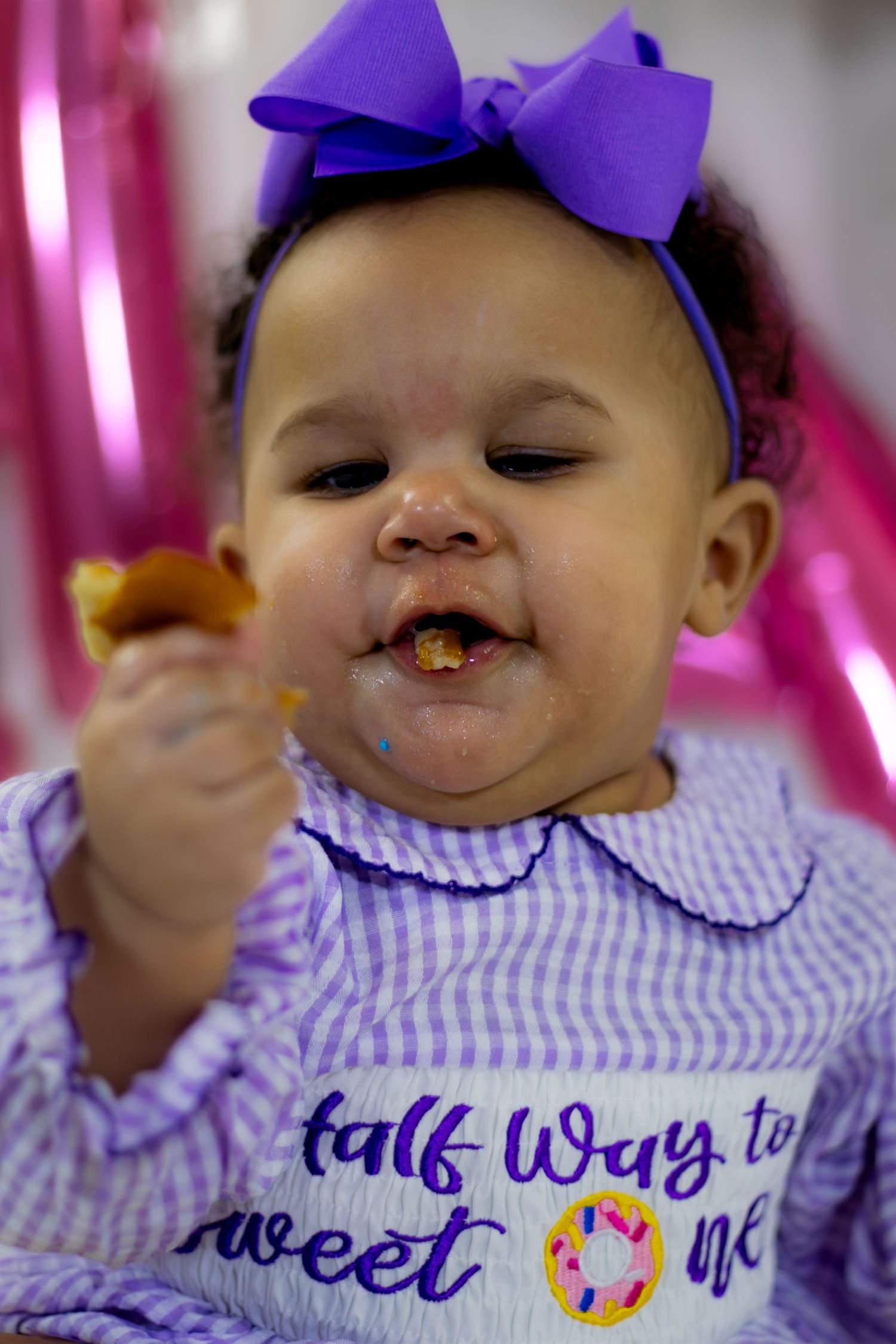 Adorable baby girl in purple gingham dress enjoying cake smash for half way to sweet one birthday.