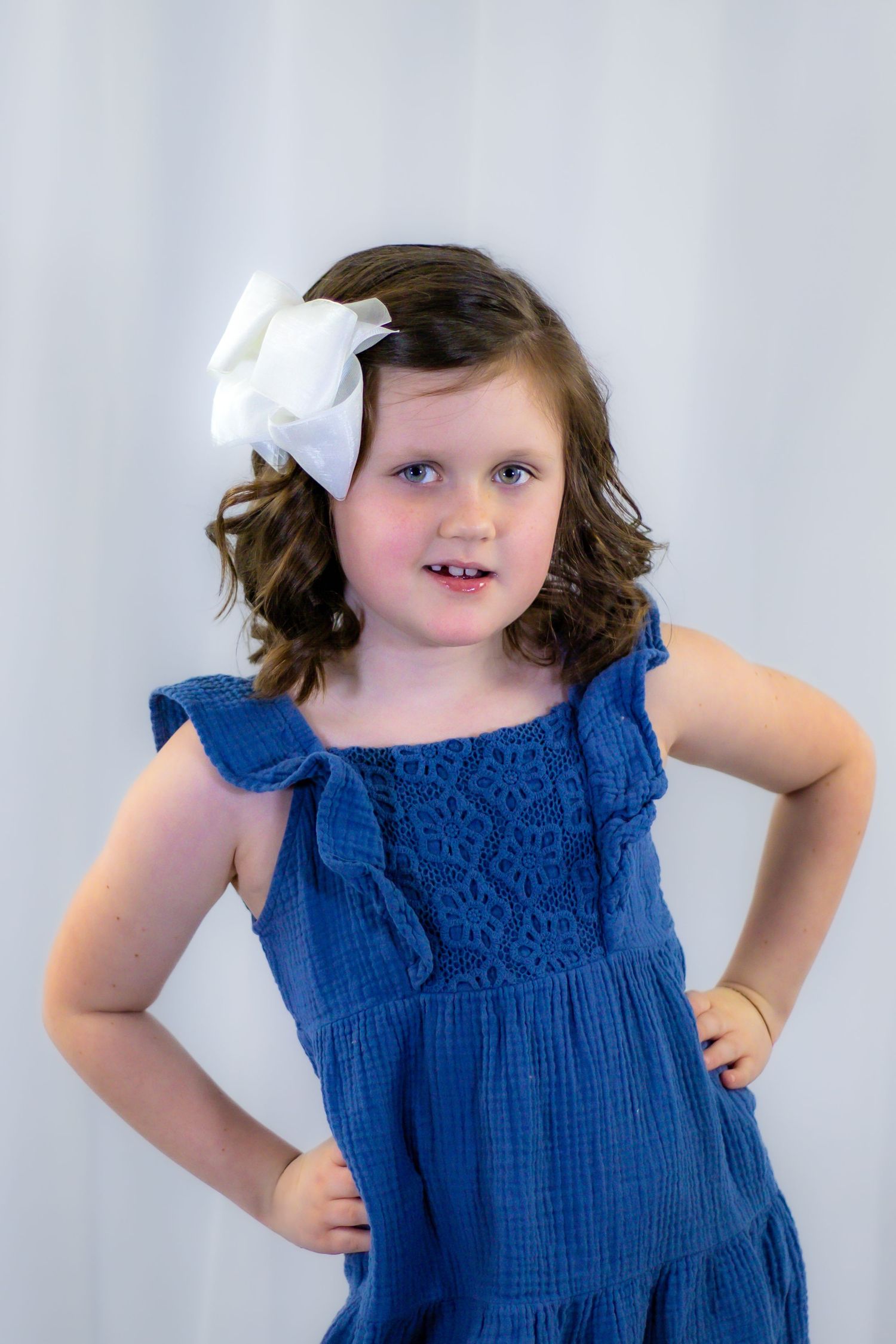 Confident young girl in blue dress with white hair bow posing hands on hips.
