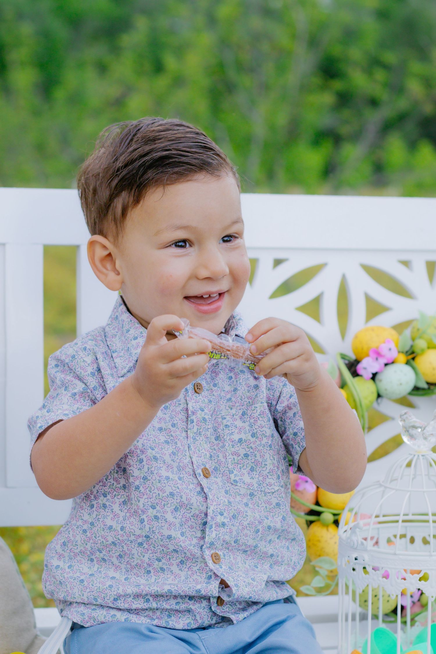 Smiling toddler boy in floral shirt sitting outdoors holding a small object near Easter decorations.
