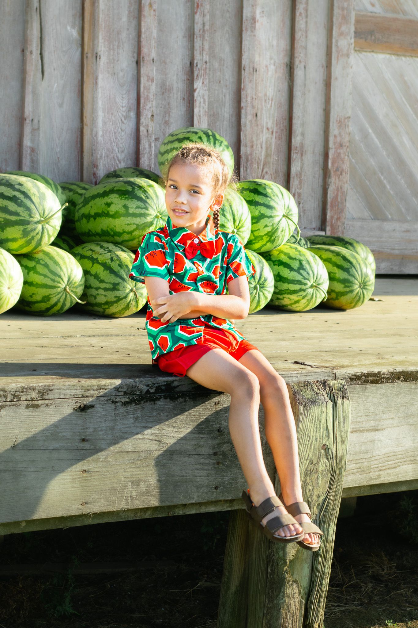 Young girl in watermelon print outfit sits on wooden platform surrounded by fresh watermelons.