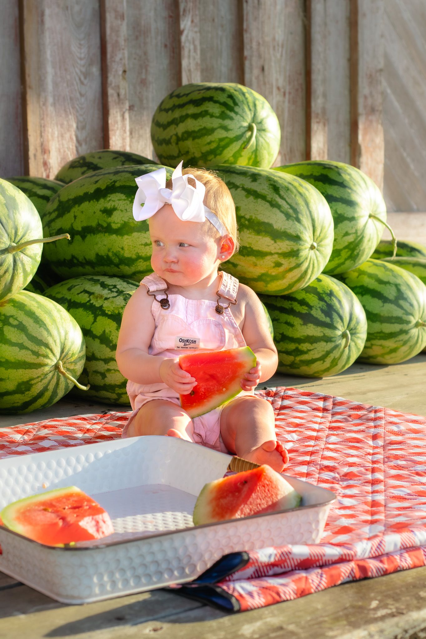 Baby girl in pink overalls and white bow sits on checkered blanket holding watermelon slice surrounded by large watermelons.