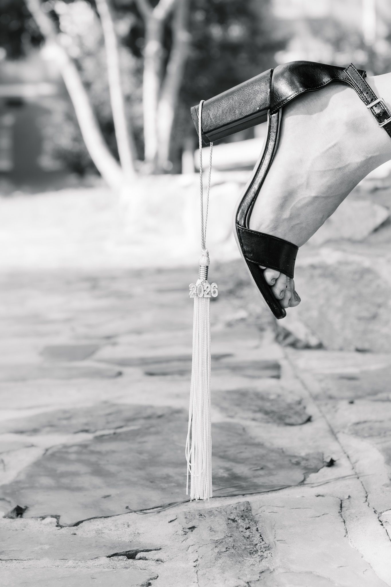 Black and white close-up of graduation tassel with 2025 charm dangling near high heel shoe.