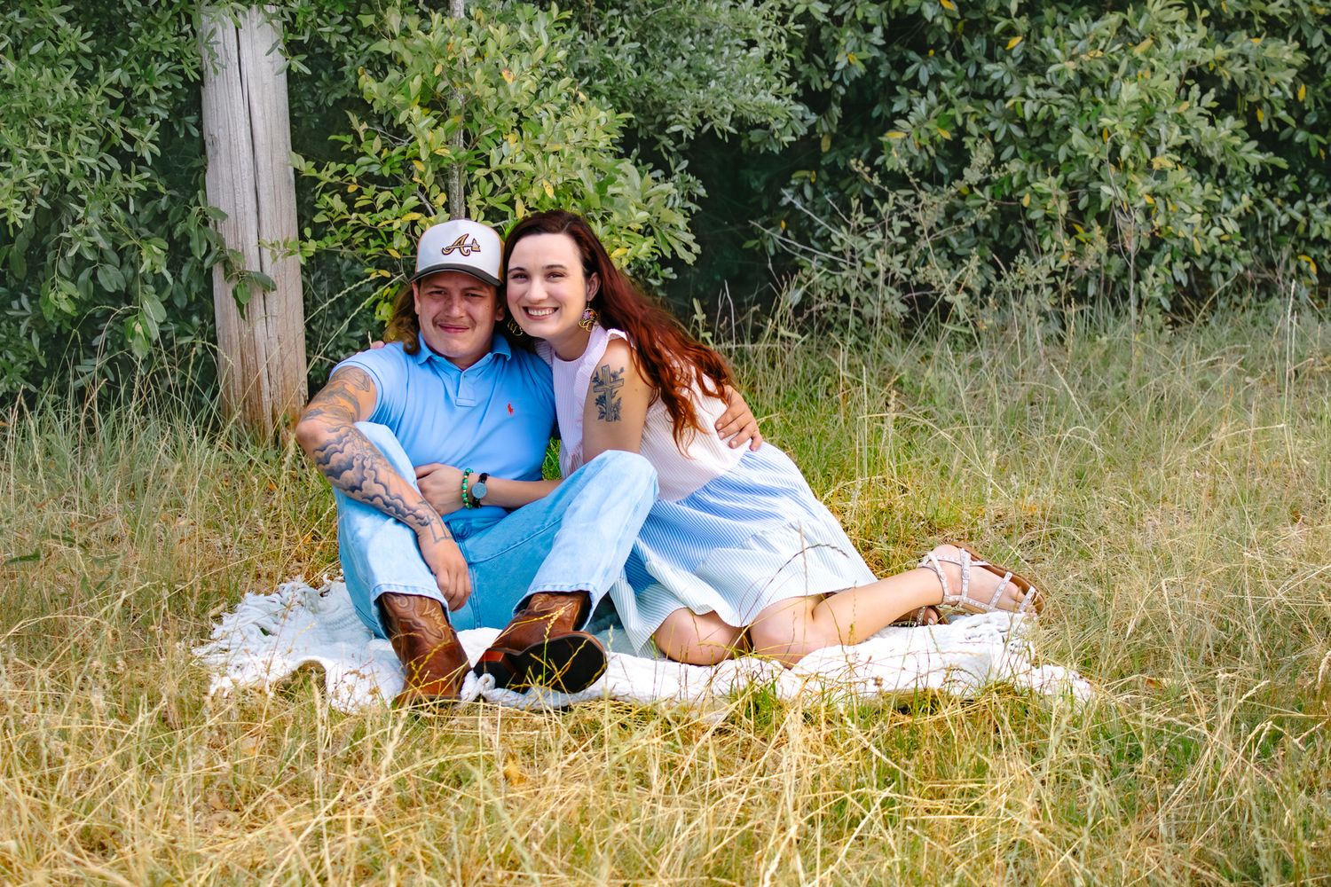 Couple sitting together on a white blanket in a grassy field smiling for a photo.