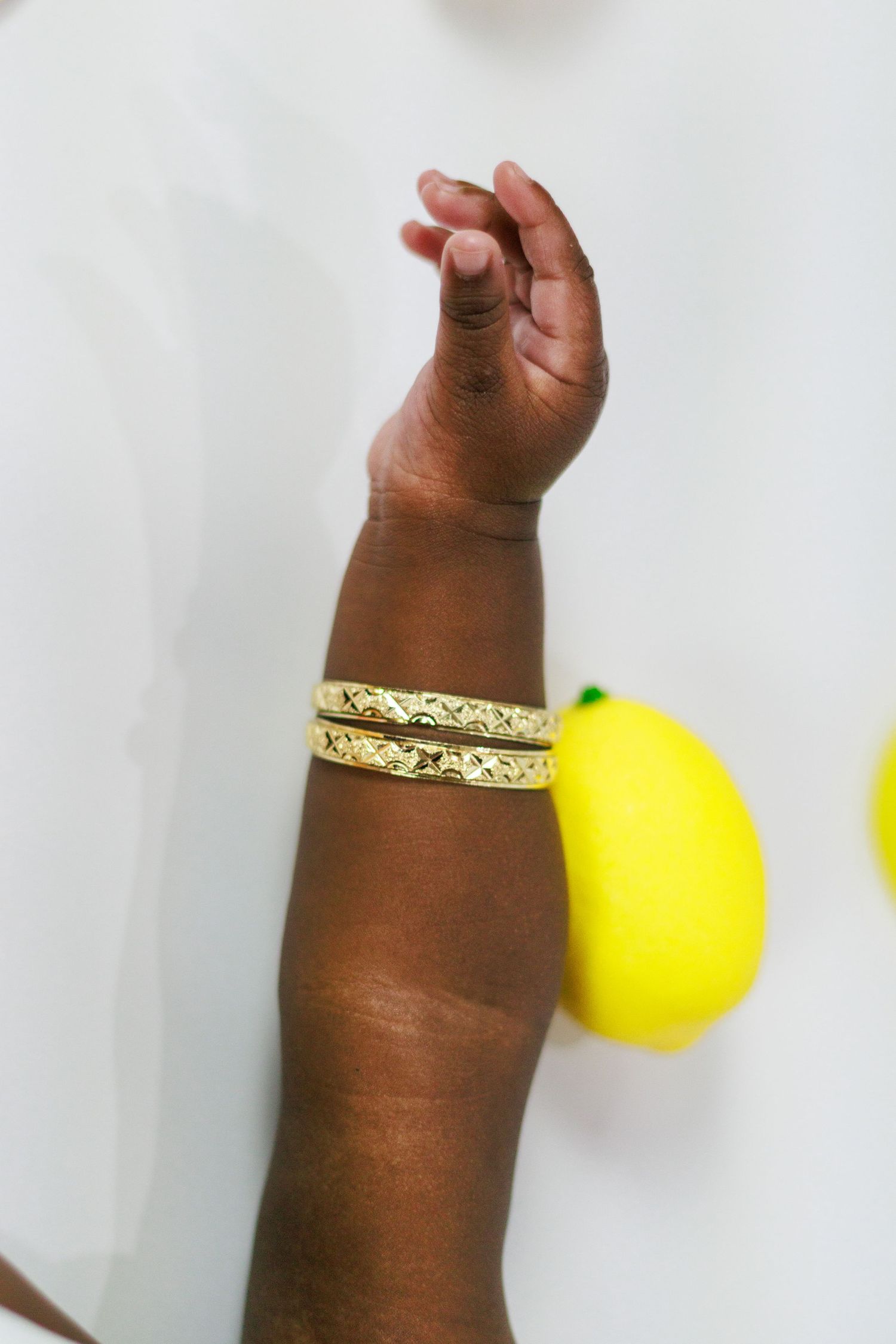 Close-up of baby hand wearing gold bangle bracelets with yellow lemon prop on white background.