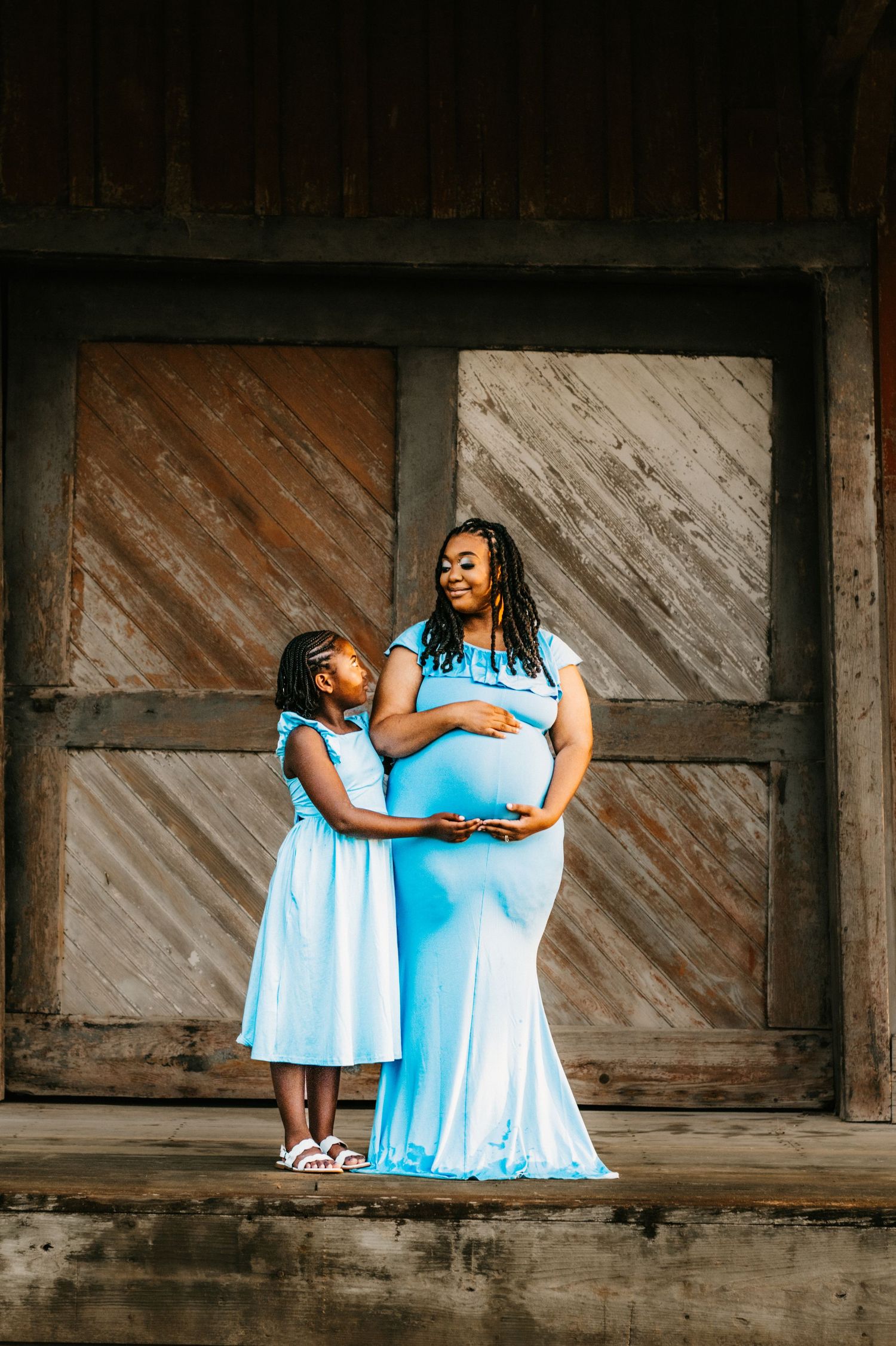 Pregnant woman in blue gown shares tender moment with young daughter touching her baby bump by barn doors.