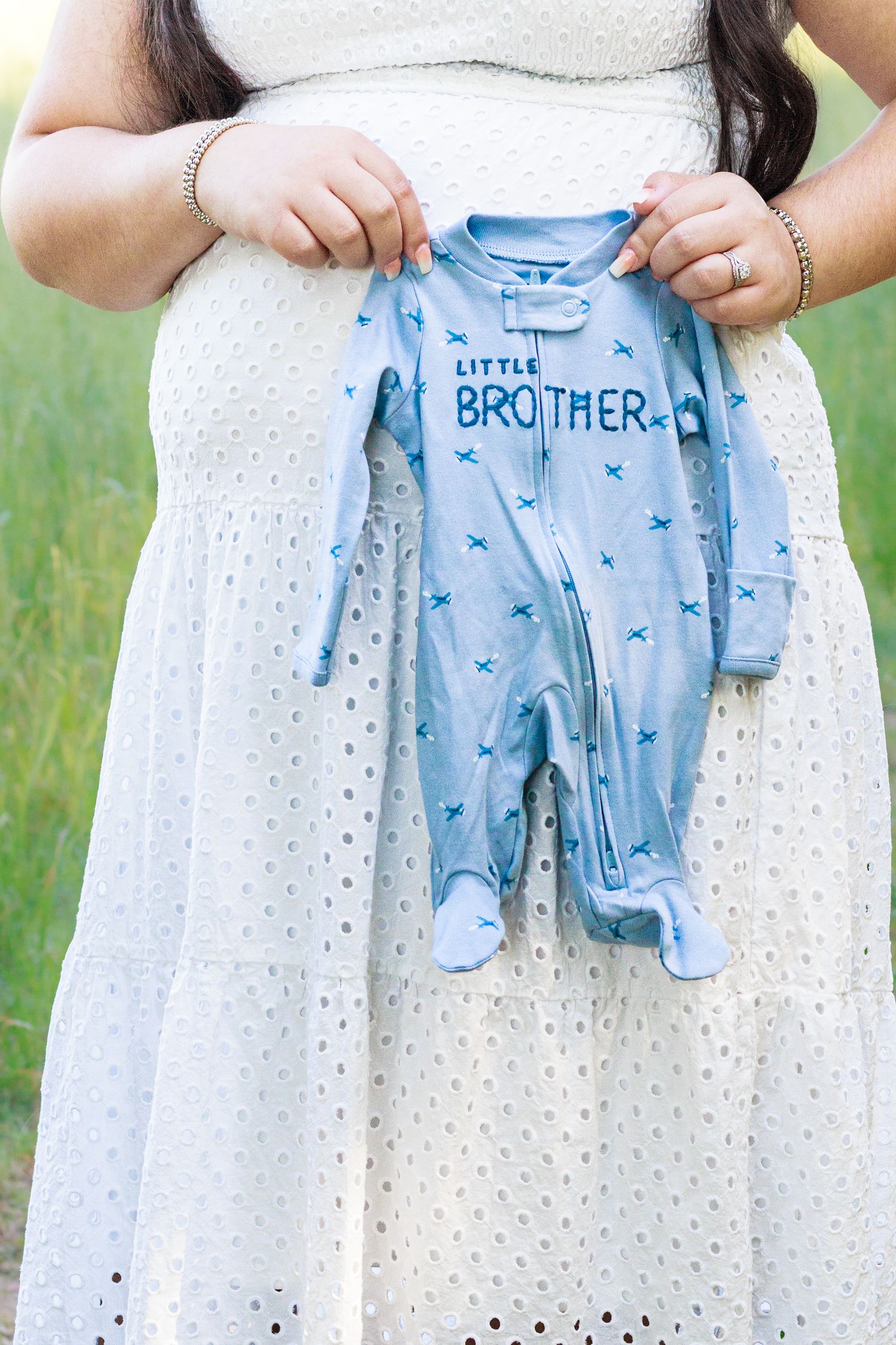 Pregnant woman in white dress holding a blue Little Brother baby onesie outdoors in a green field.