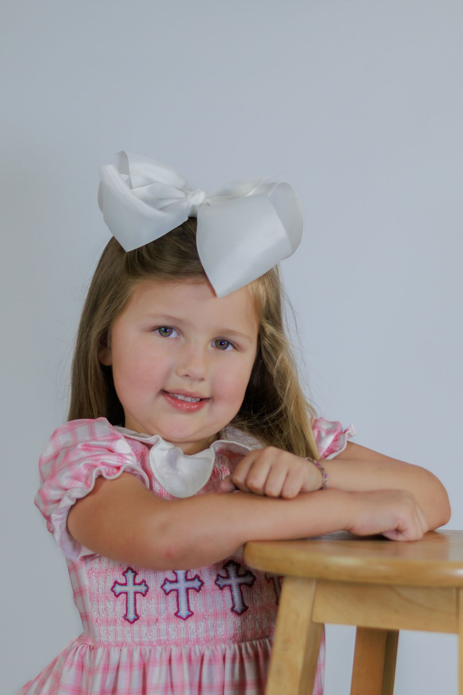 Sweet little girl wearing pink smocked dress with cross details and large white bow, leaning on wooden stool.