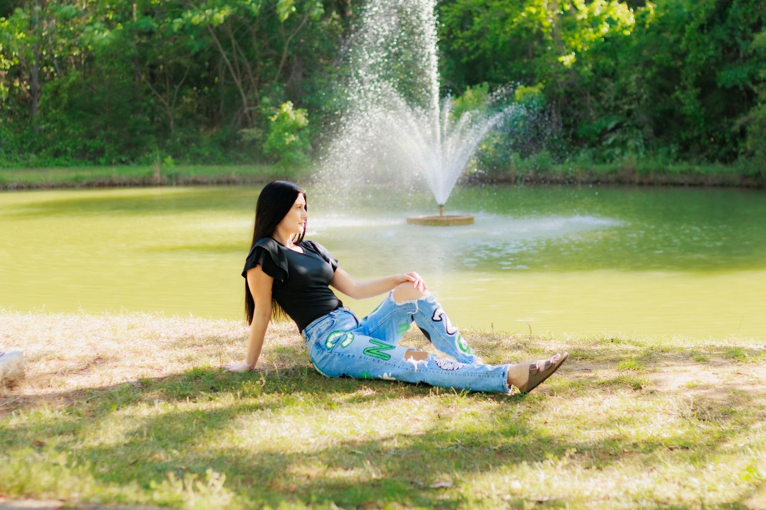 Young woman in ripped jeans relaxing on grass by a pond with a fountain in a lush green park.