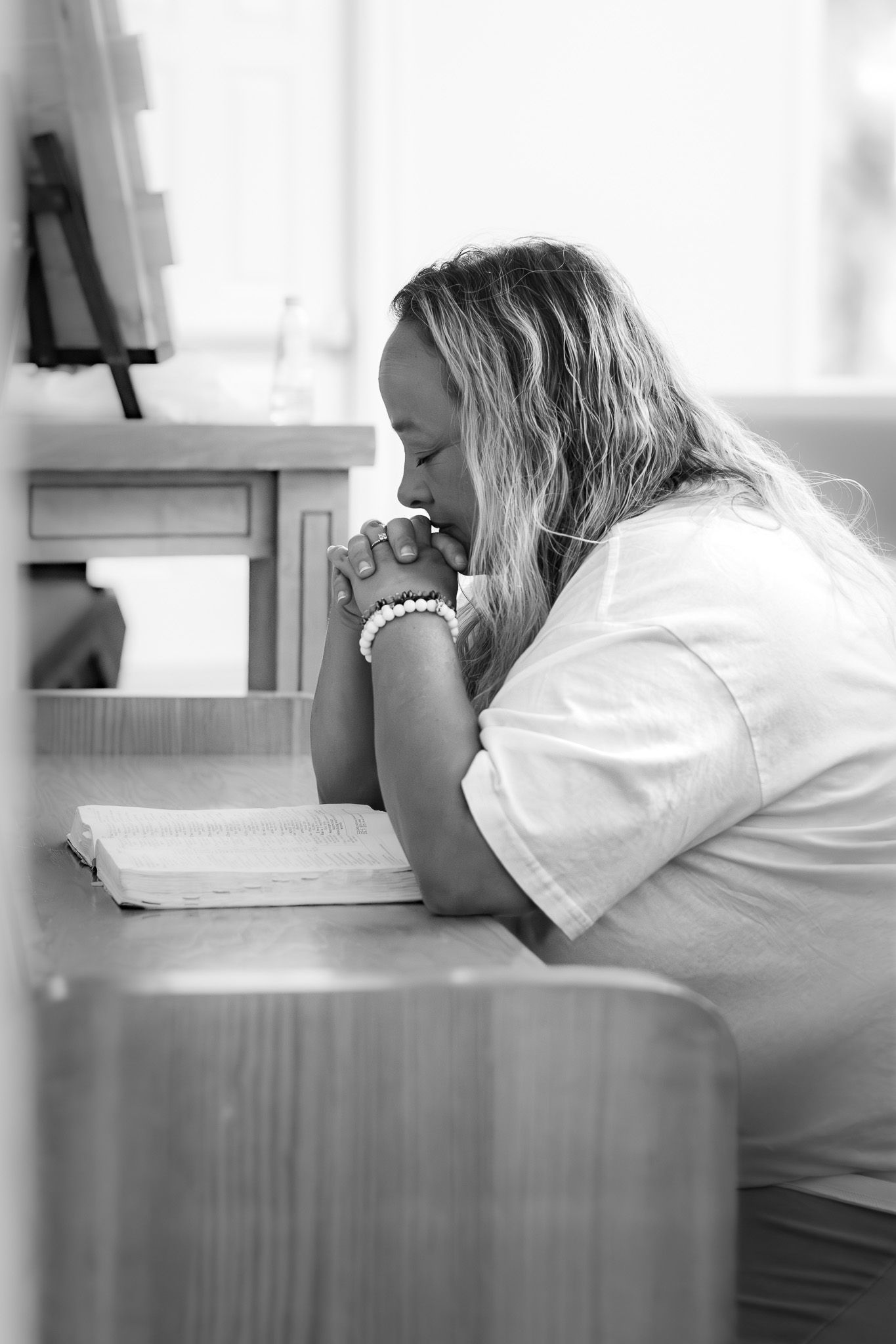 Black and white photo of a woman praying with clasped hands over an open Bible at a wooden desk.
