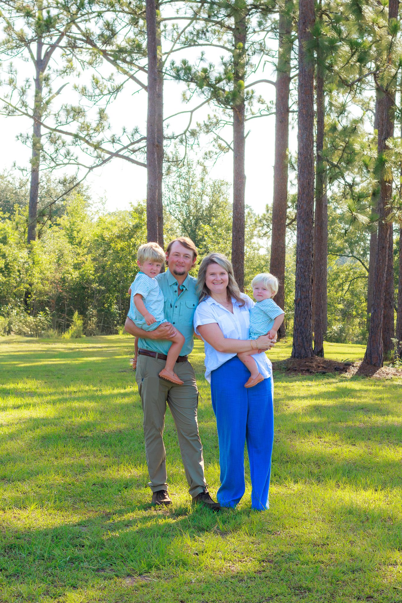 Family of four poses outdoors among tall pine trees, parents holding two young boys in matching teal outfits on green lawn.