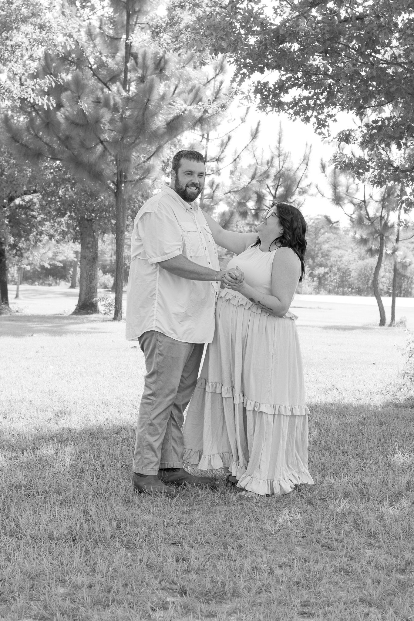 Black and white photo of a pregnant couple standing in a park, smiling and holding hands among lush trees.
