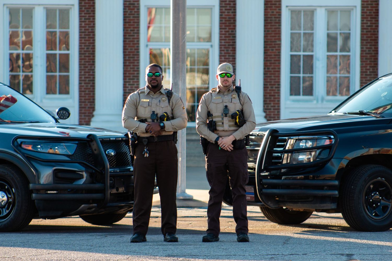 Two sheriff deputies in uniform stand between patrol vehicles in front of a government building.