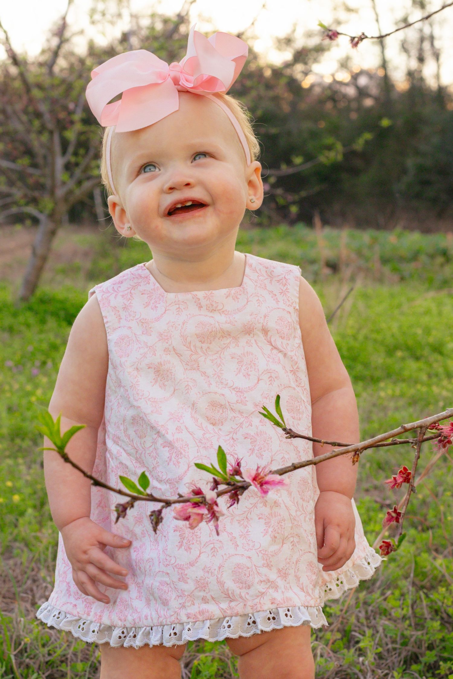 Adorable smiling baby girl in pink paisley dress holding blooming spring branch in outdoor orchard setting.