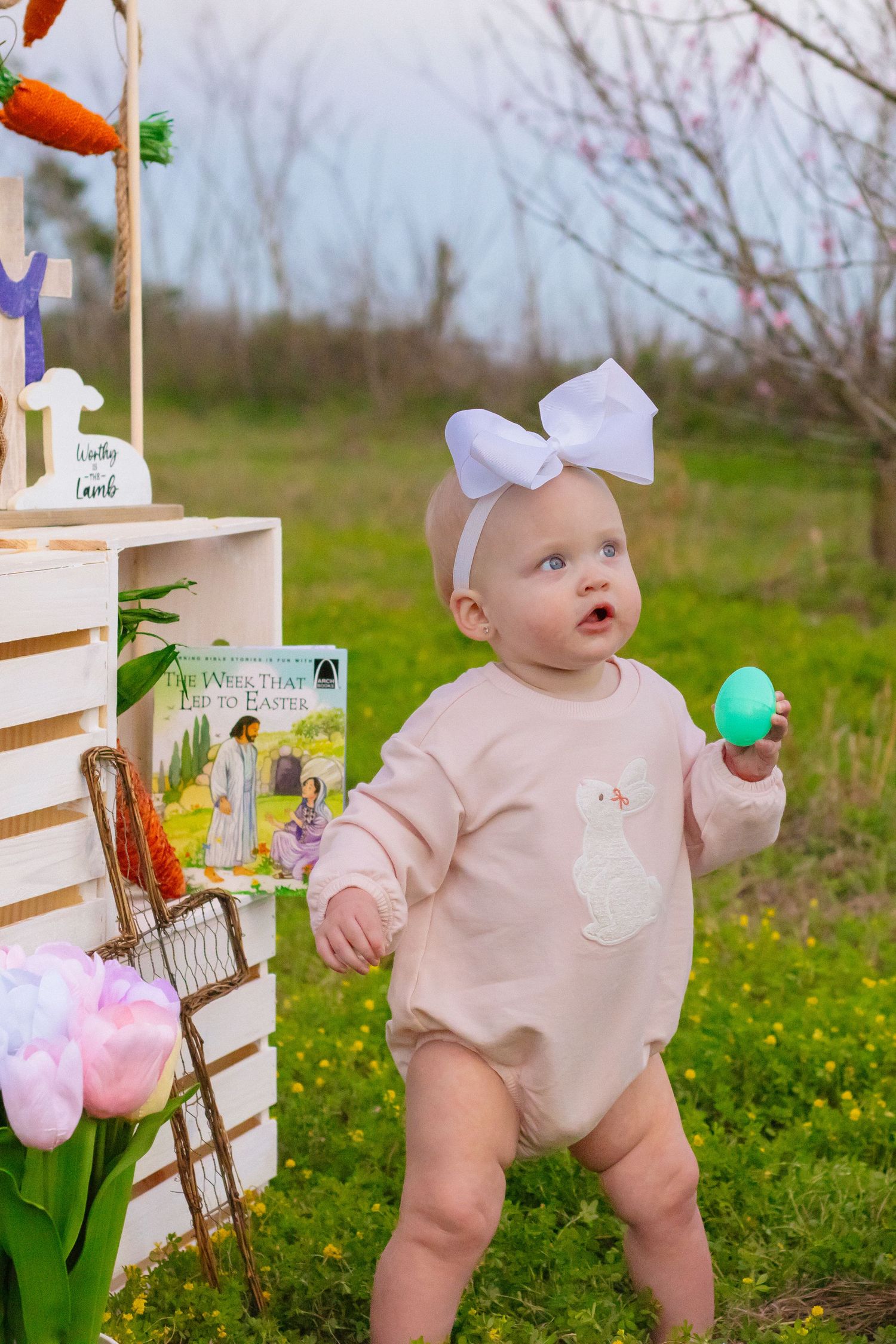 Baby in pink romper with white bow holds green Easter egg standing in spring field.