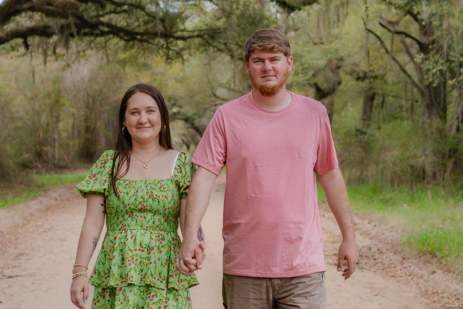 Couple holding hands walking down a scenic dirt path surrounded by lush green trees.