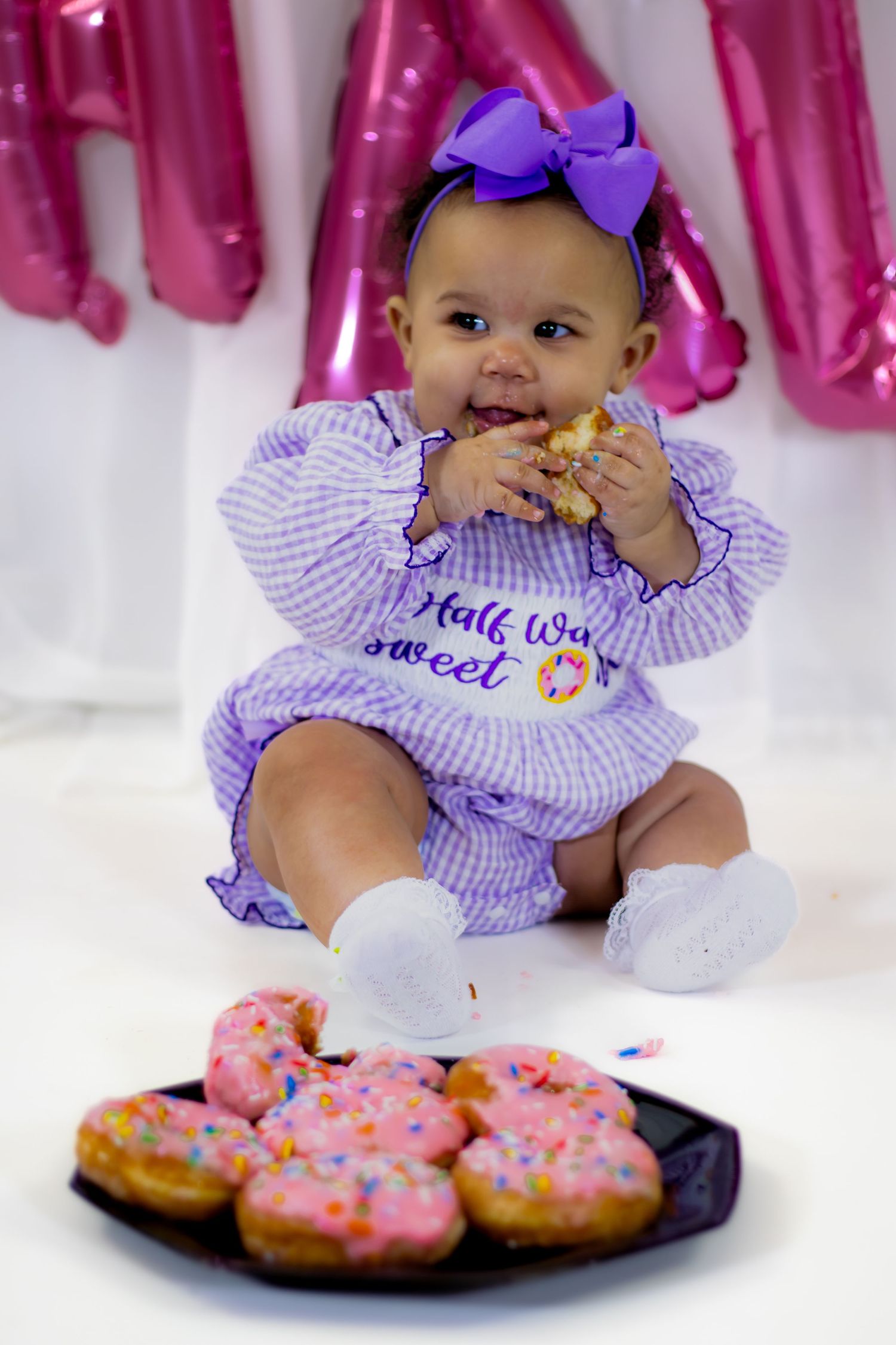 Cute baby in purple gingham outfit eating donut beside pink frosted donuts at celebration.