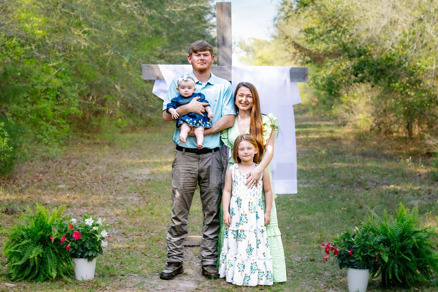 Young family of four posing before a wooden cross with potted ferns in an outdoor setting.