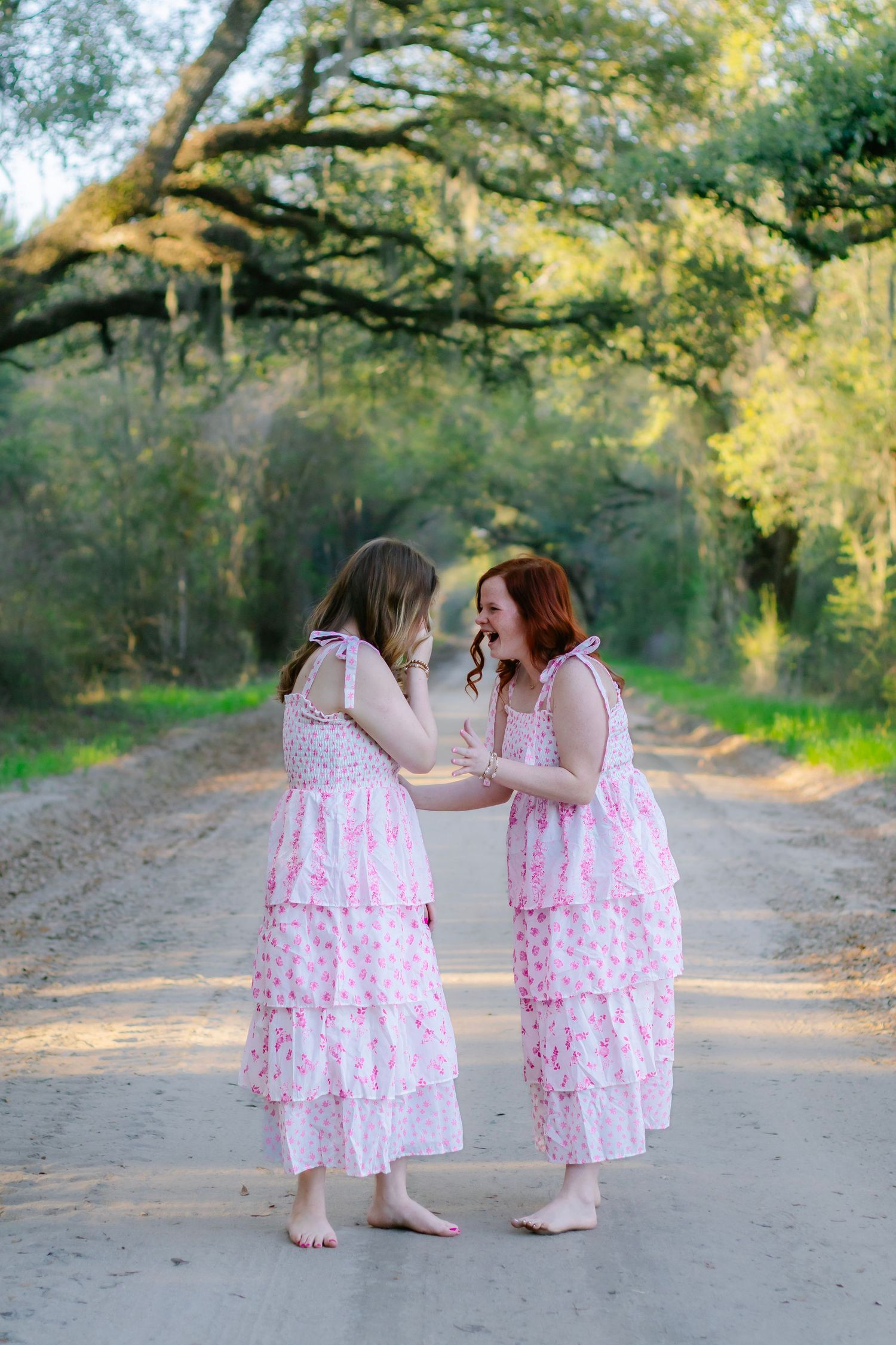 Two barefoot girls in matching pink tiered dresses laugh together on a tree-lined dirt road.
