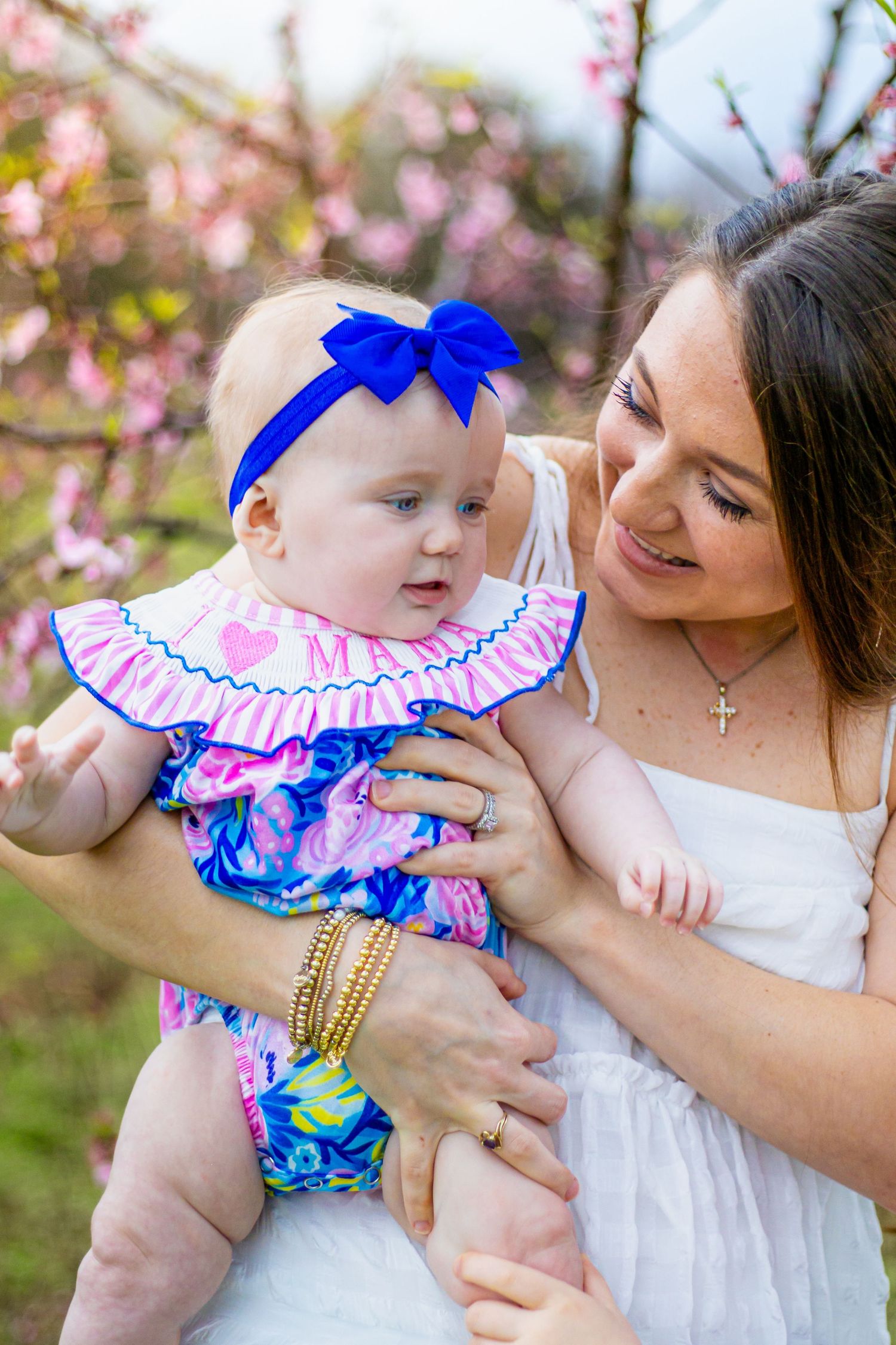 Mother in white dress lovingly holds baby girl in pink floral outfit with blue bow headband.