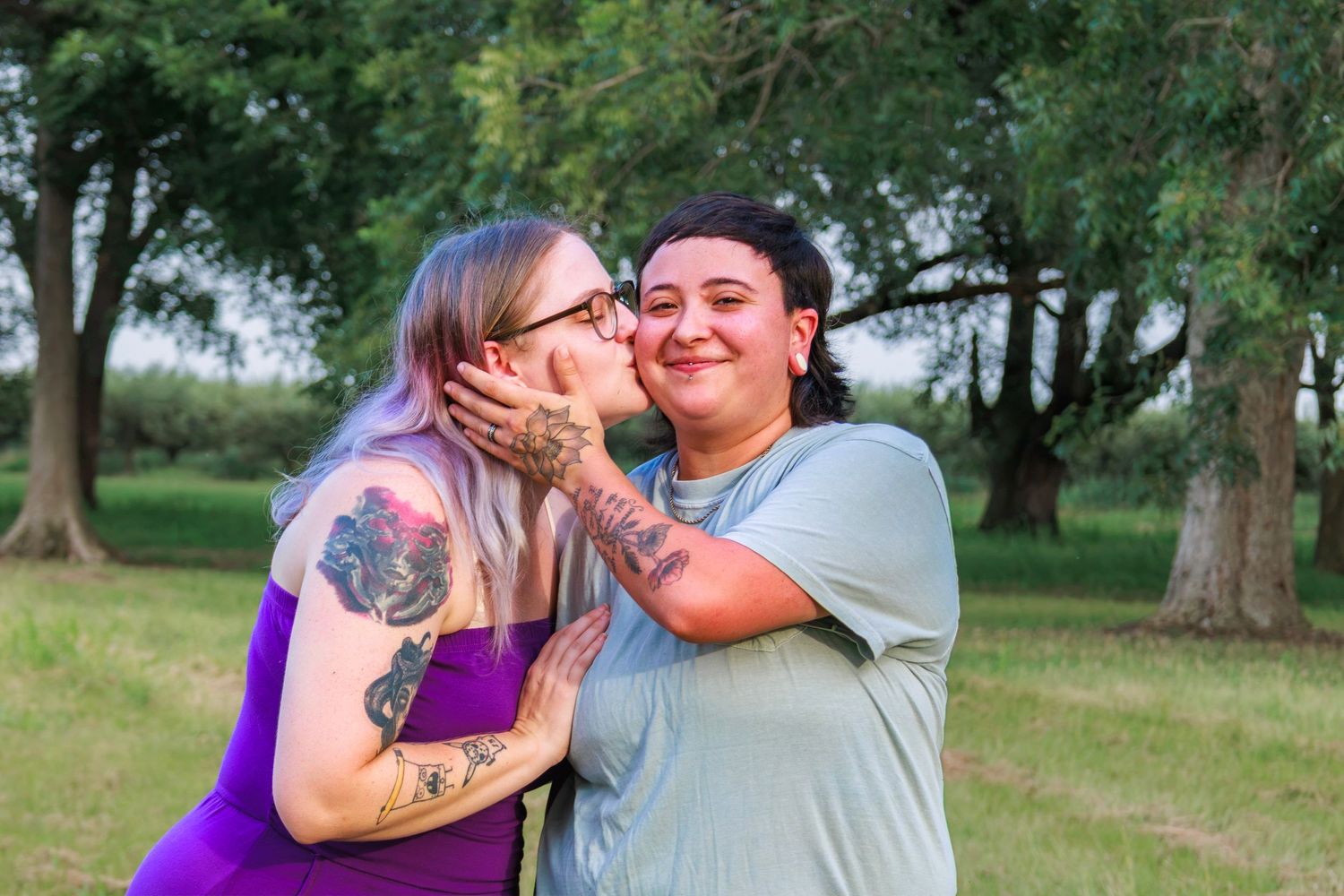 Two women share a sweet kiss outdoors, one with tattoos in a purple dress, surrounded by lush green trees.