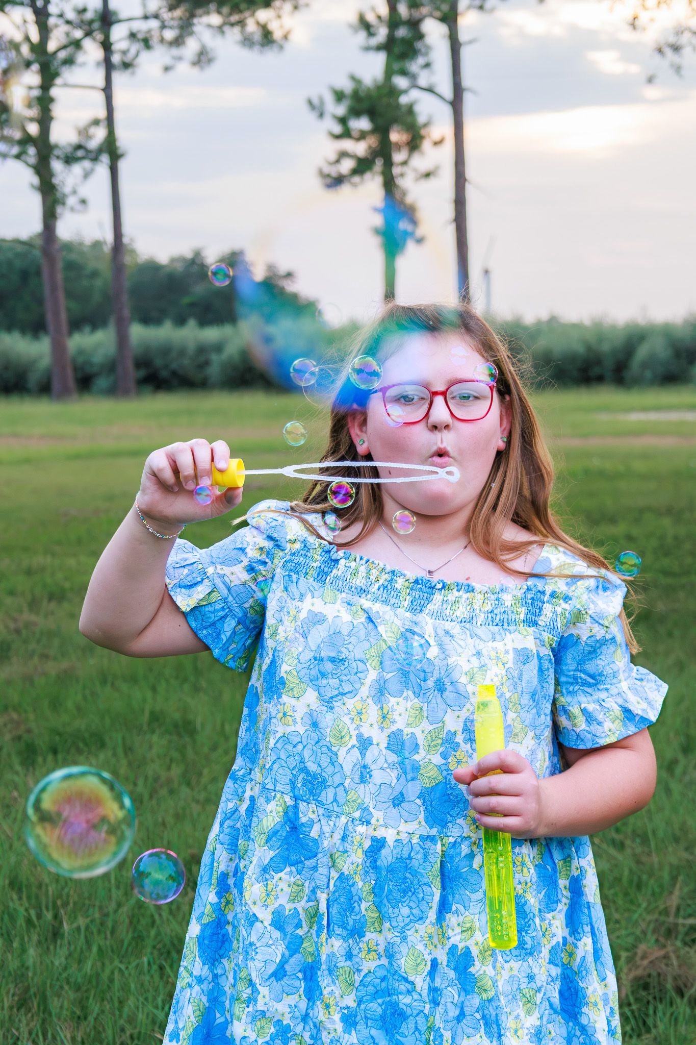 A young girl with red glasses blows soap bubbles outdoors in a blue floral dress, holding bubble wands in a green park.