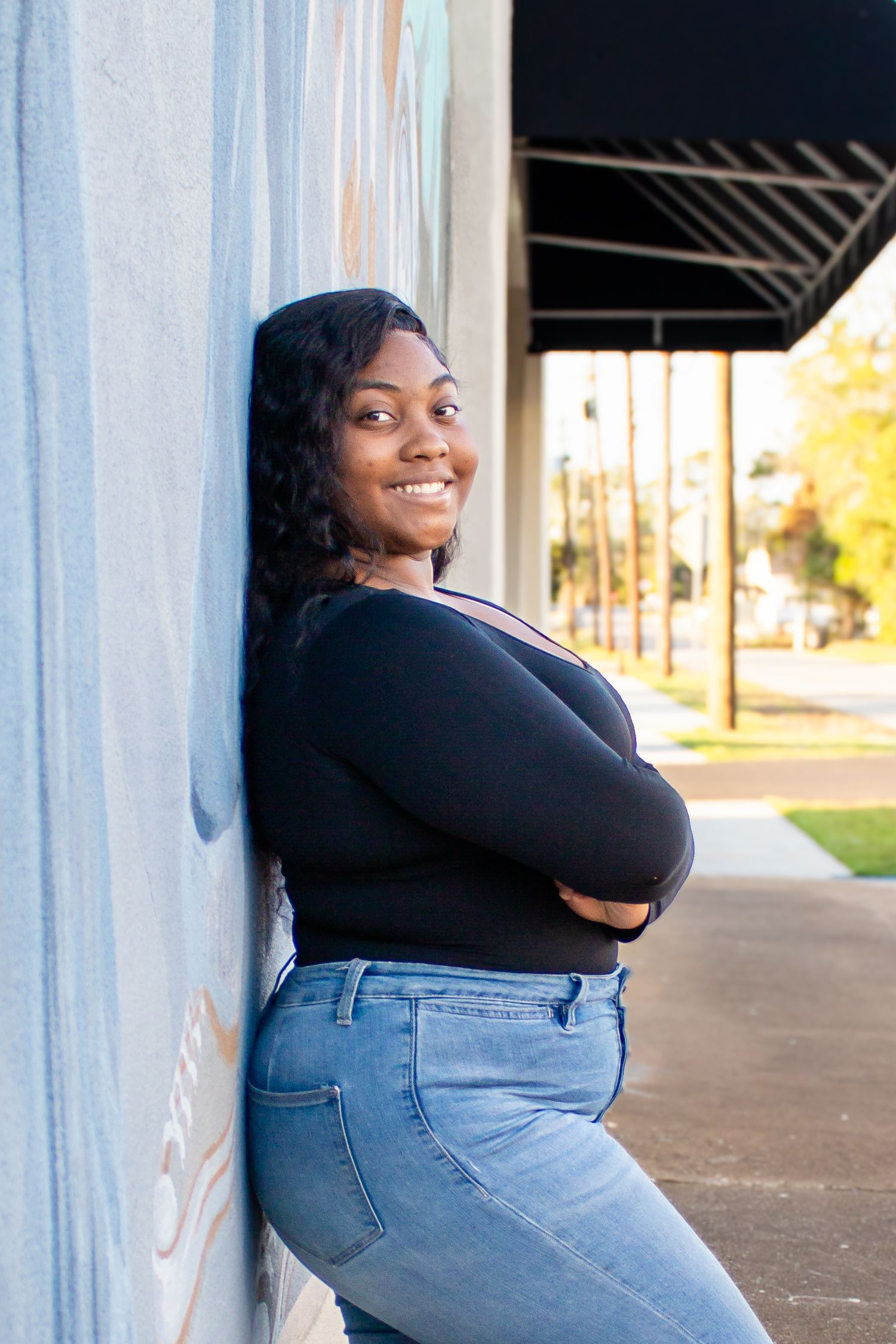 Young Black woman in black top and jeans leaning against colorful mural wall with arms crossed.