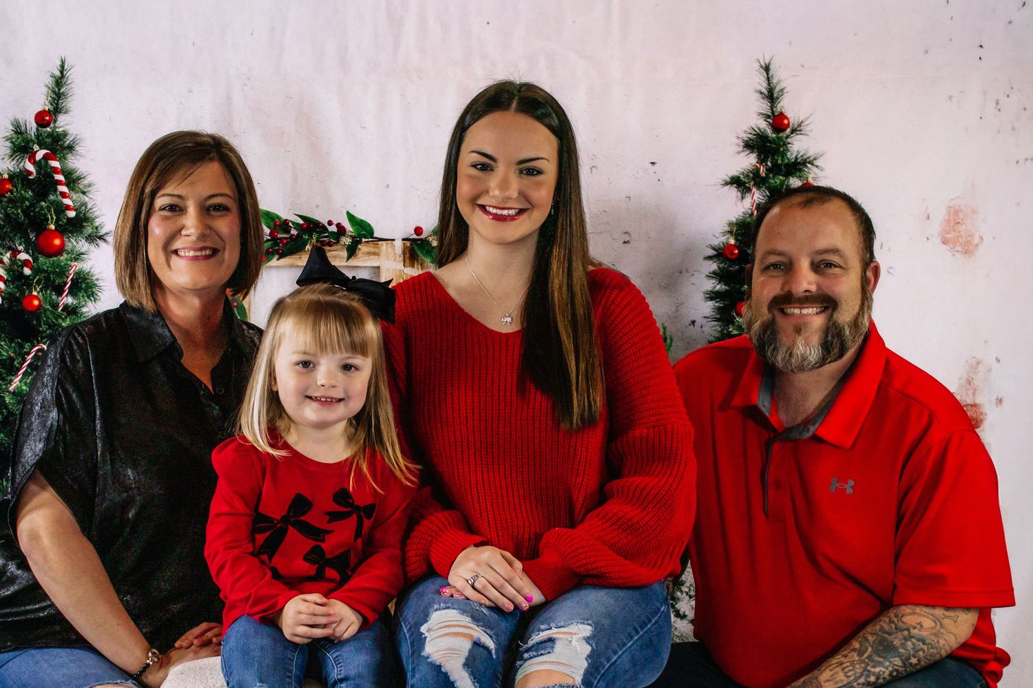 Family of four smiling together in front of Christmas trees for holiday portrait session.
