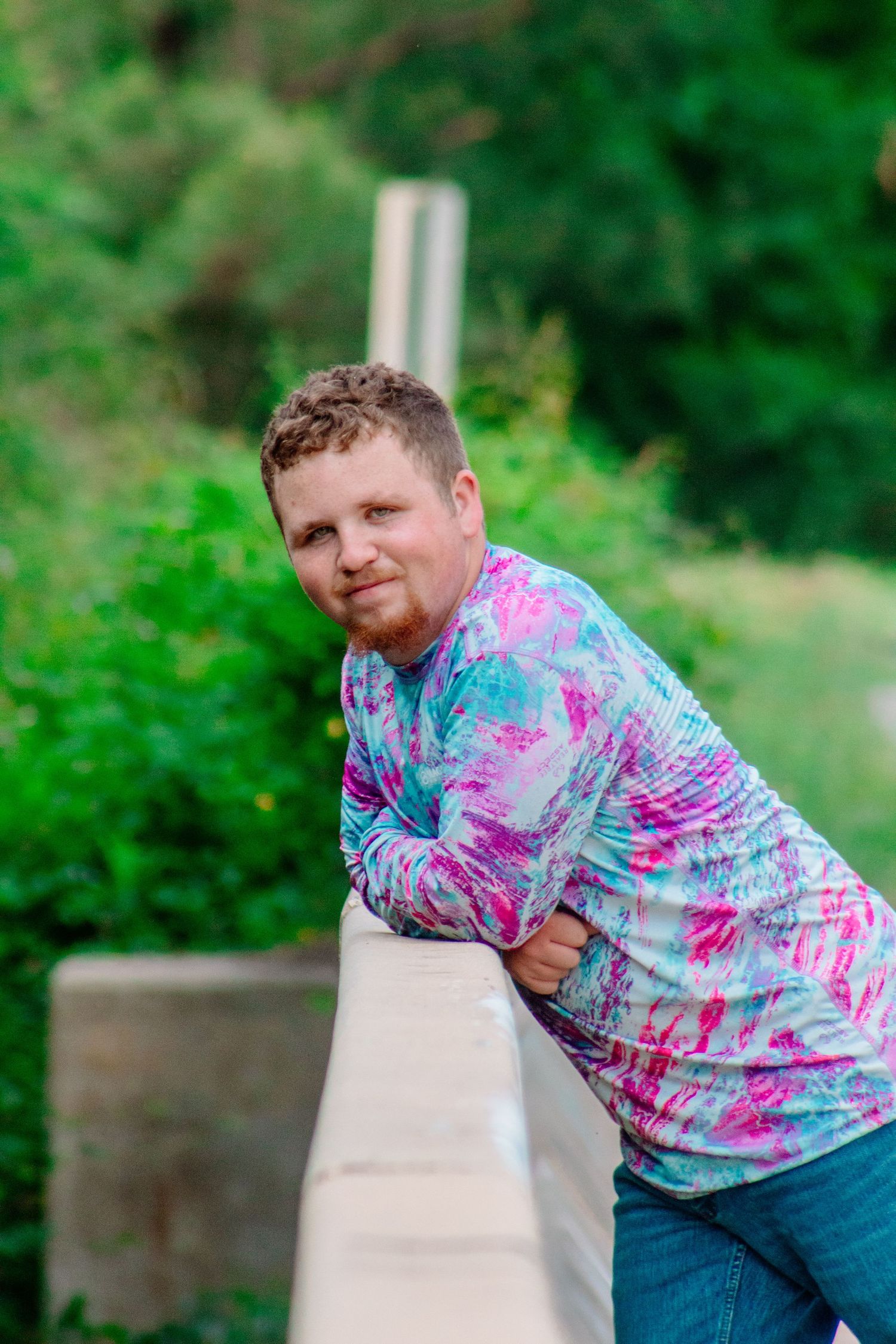 Man in colorful tie-dye shirt leaning on a bridge railing surrounded by lush greenery.