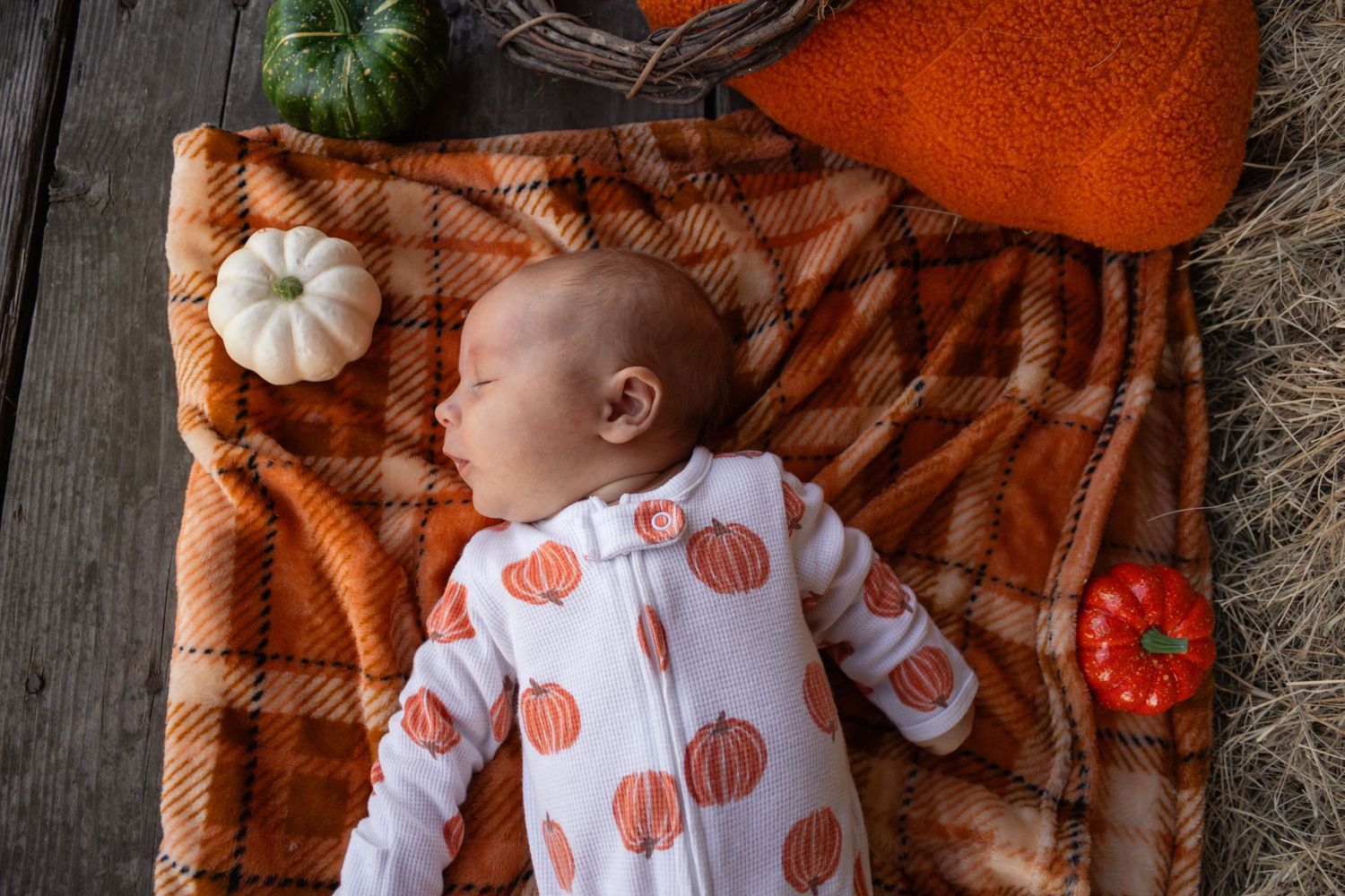 Newborn baby in pumpkin-print pajamas lies on an orange plaid blanket surrounded by small pumpkins.