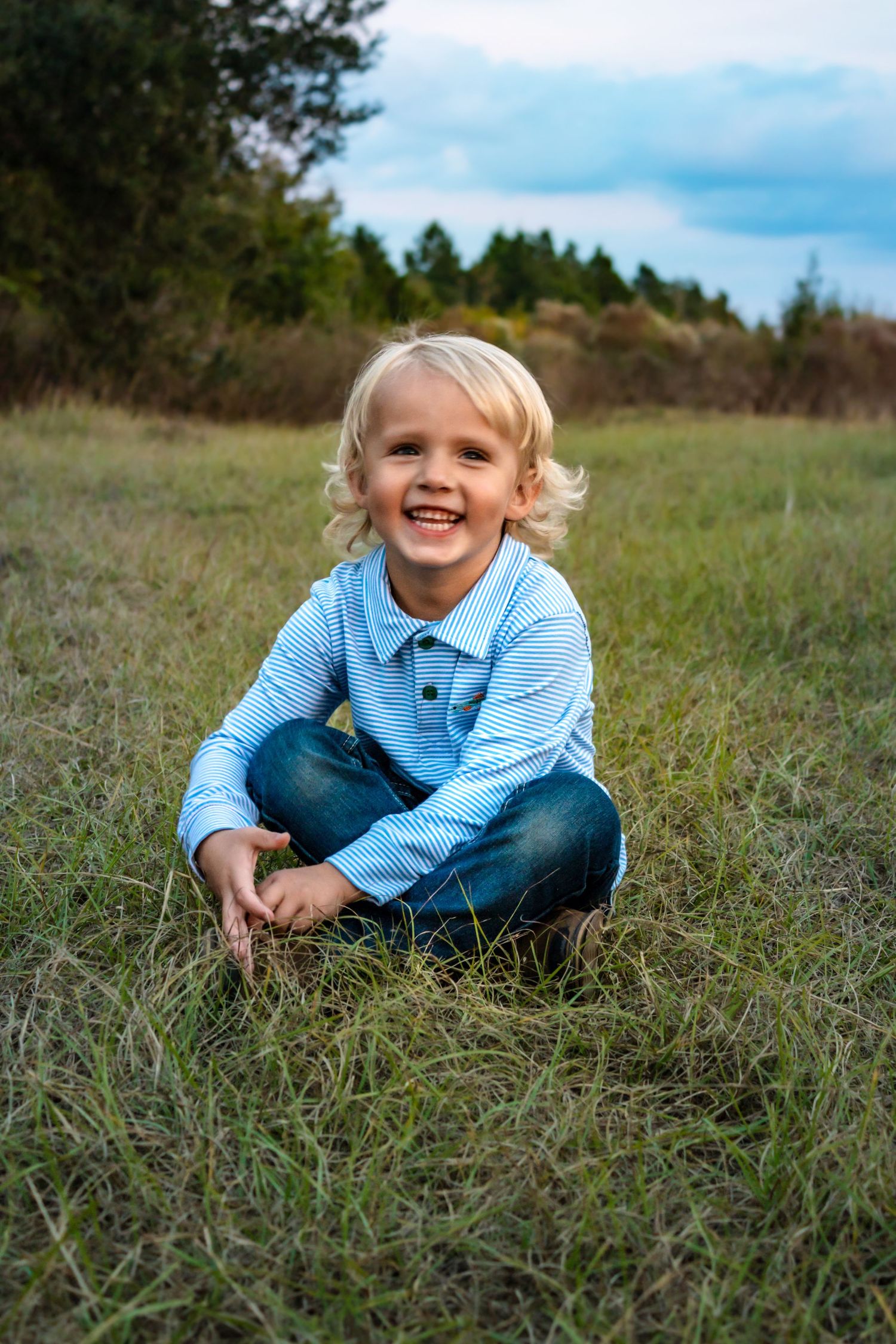 Laughing blonde toddler boy in blue striped shirt sitting barefoot on green grass in open field.