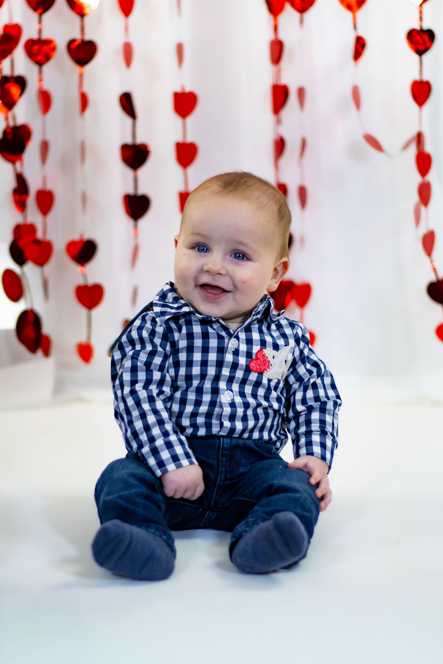 Happy baby in blue gingham shirt and jeans sits smiling before red heart garland backdrop.