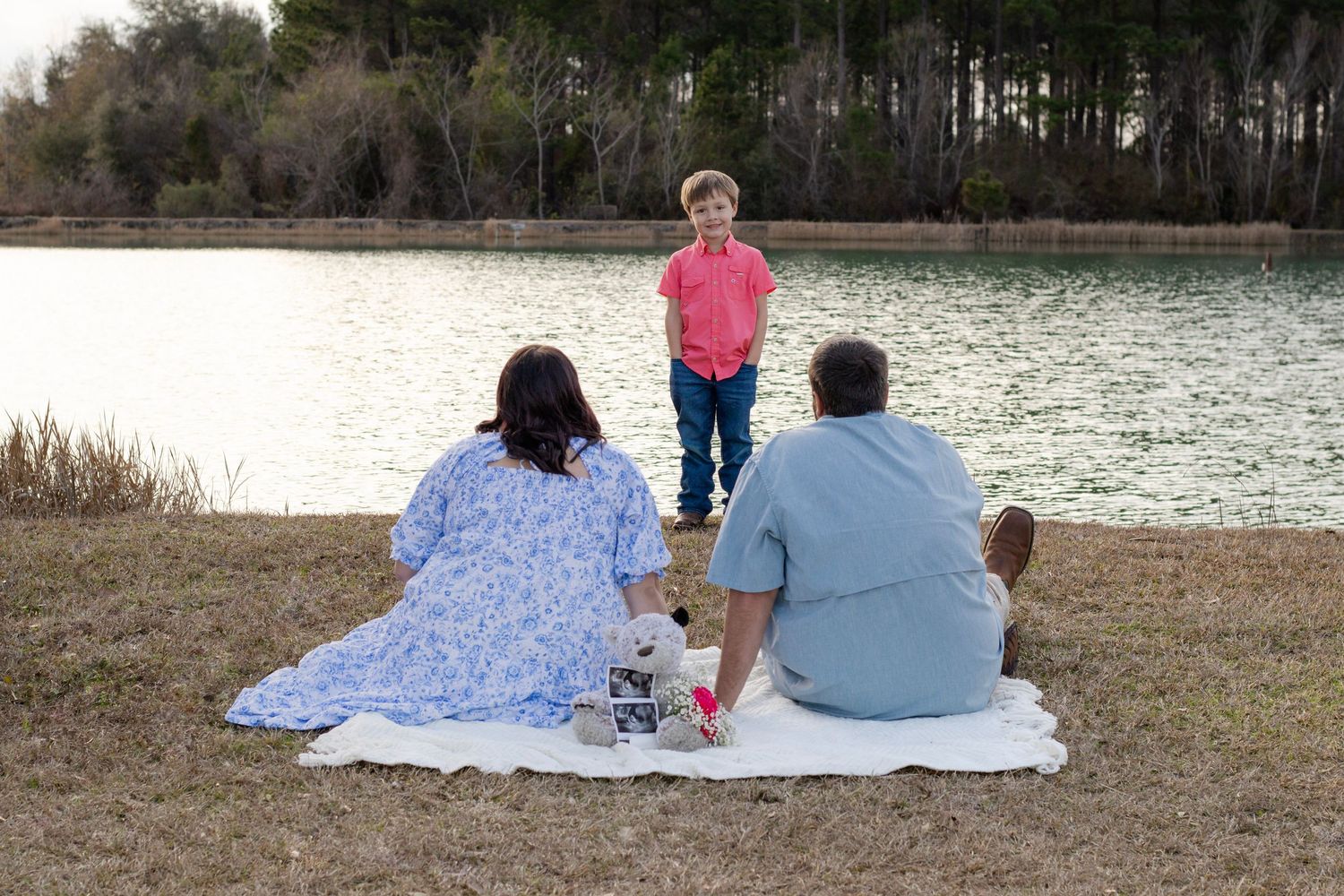 Pregnant couple sits on blanket by lake as young boy stands before them near ultrasound photos.