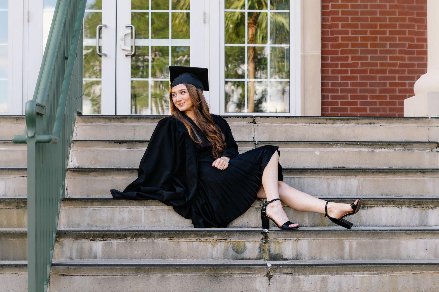 Graduate in cap and gown sitting relaxed on outdoor building steps.