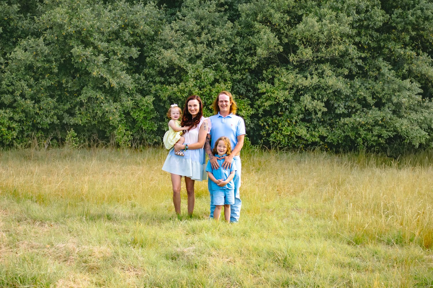 Two women and two young children smiling outdoors in a green meadow with trees in the background.