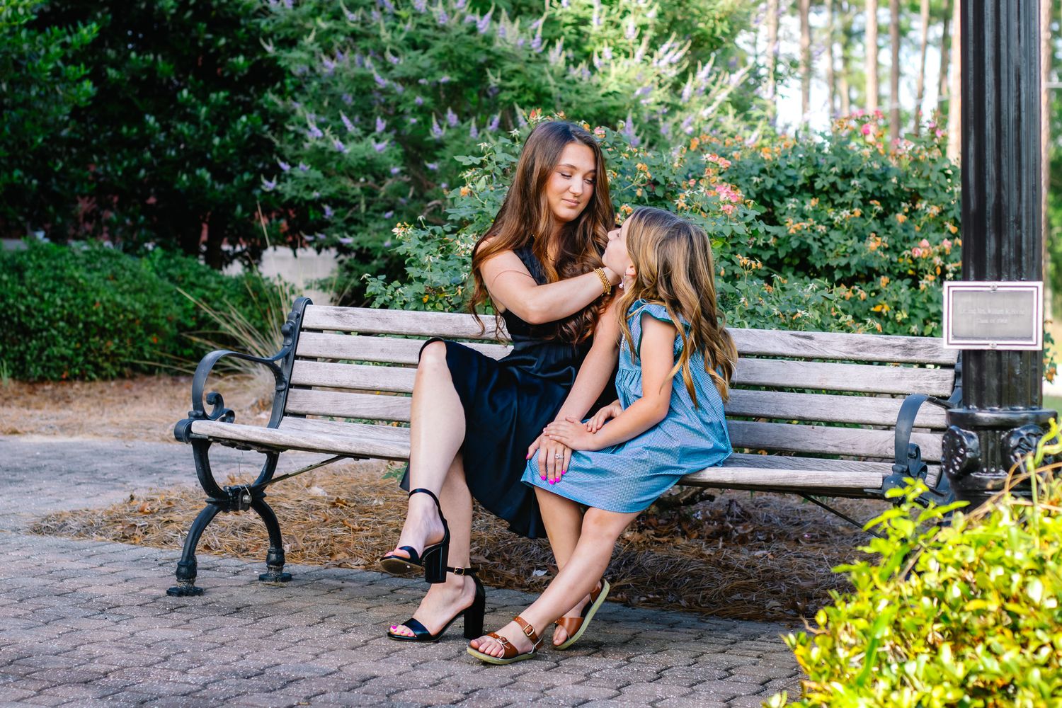 A mother tenderly fixes her young daughter's hair while sitting together on a park bench outdoors.