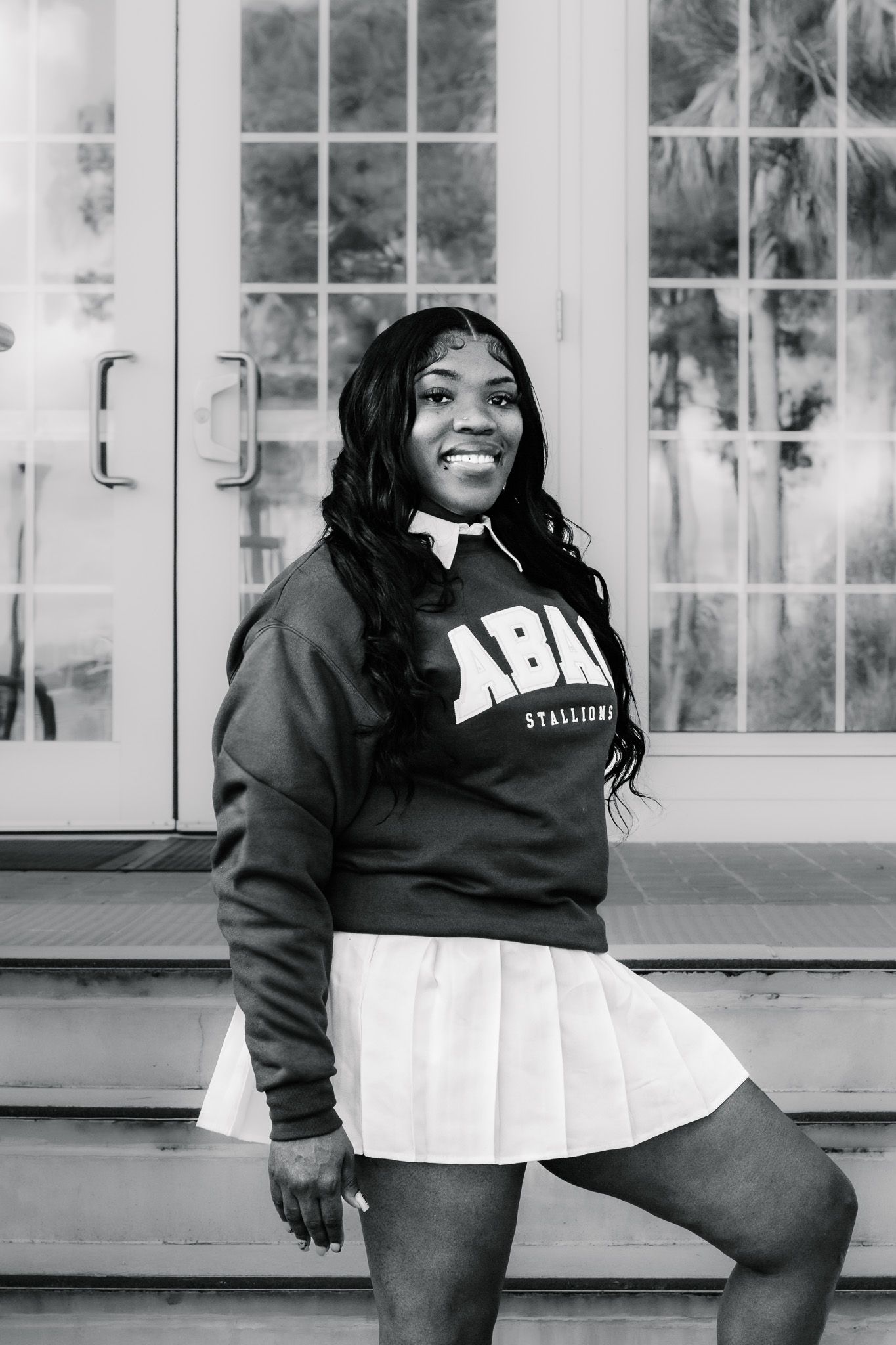 Smiling young woman in ABAI Stallions sweatshirt and white skirt posing outdoors in black and white photo.
