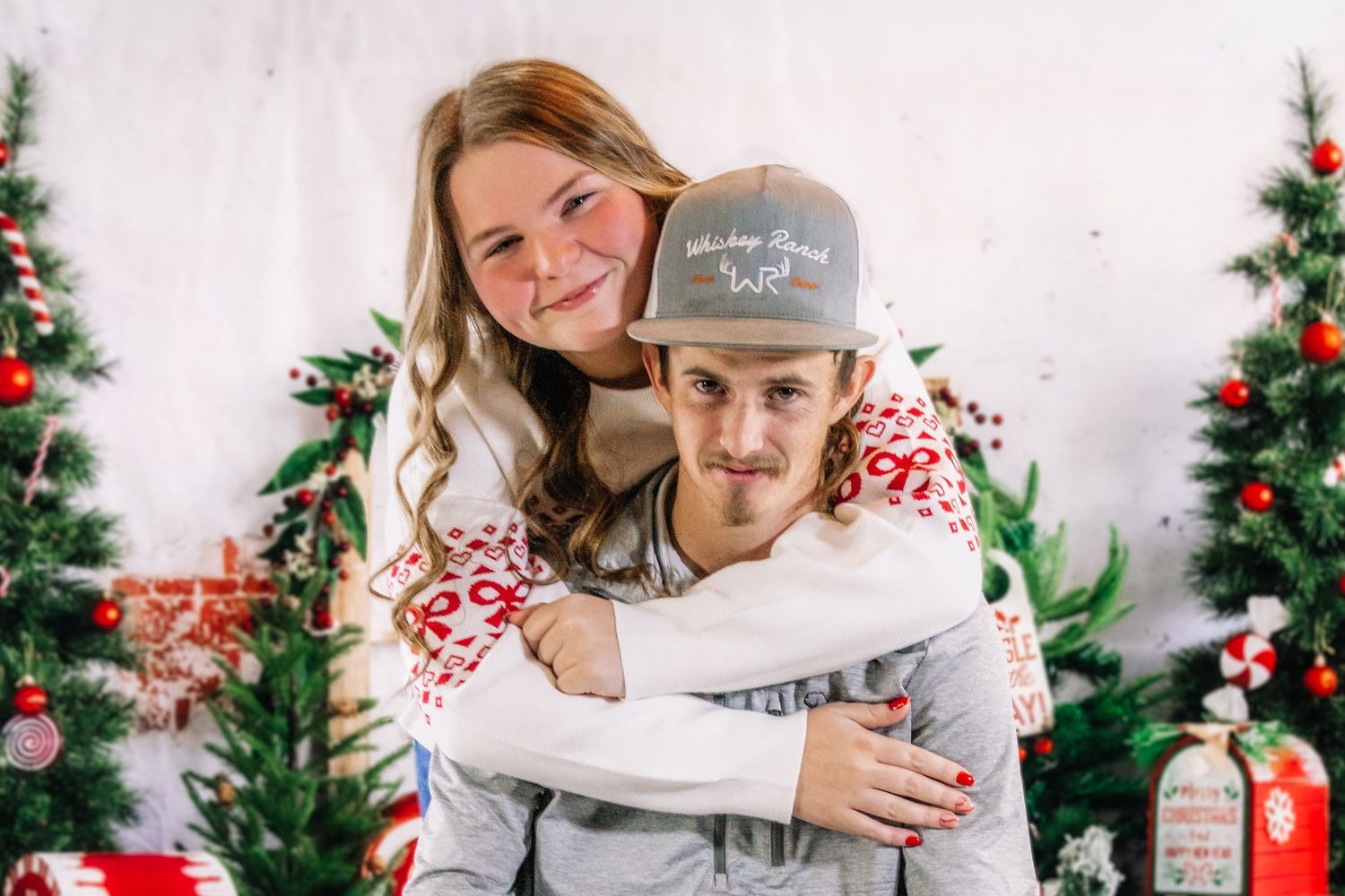 A young couple in matching holiday sweaters embrace playfully in front of decorated Christmas trees in a studio.