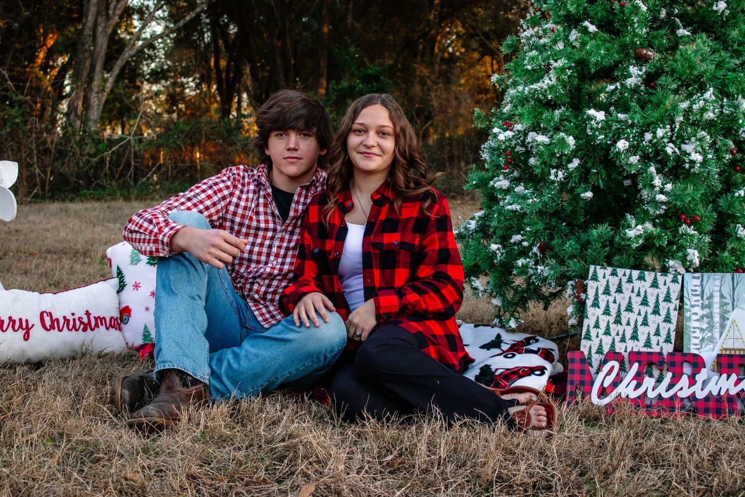 Young couple in red plaid shirts sitting together on a blanket beside a decorated outdoor Christmas tree at sunset.